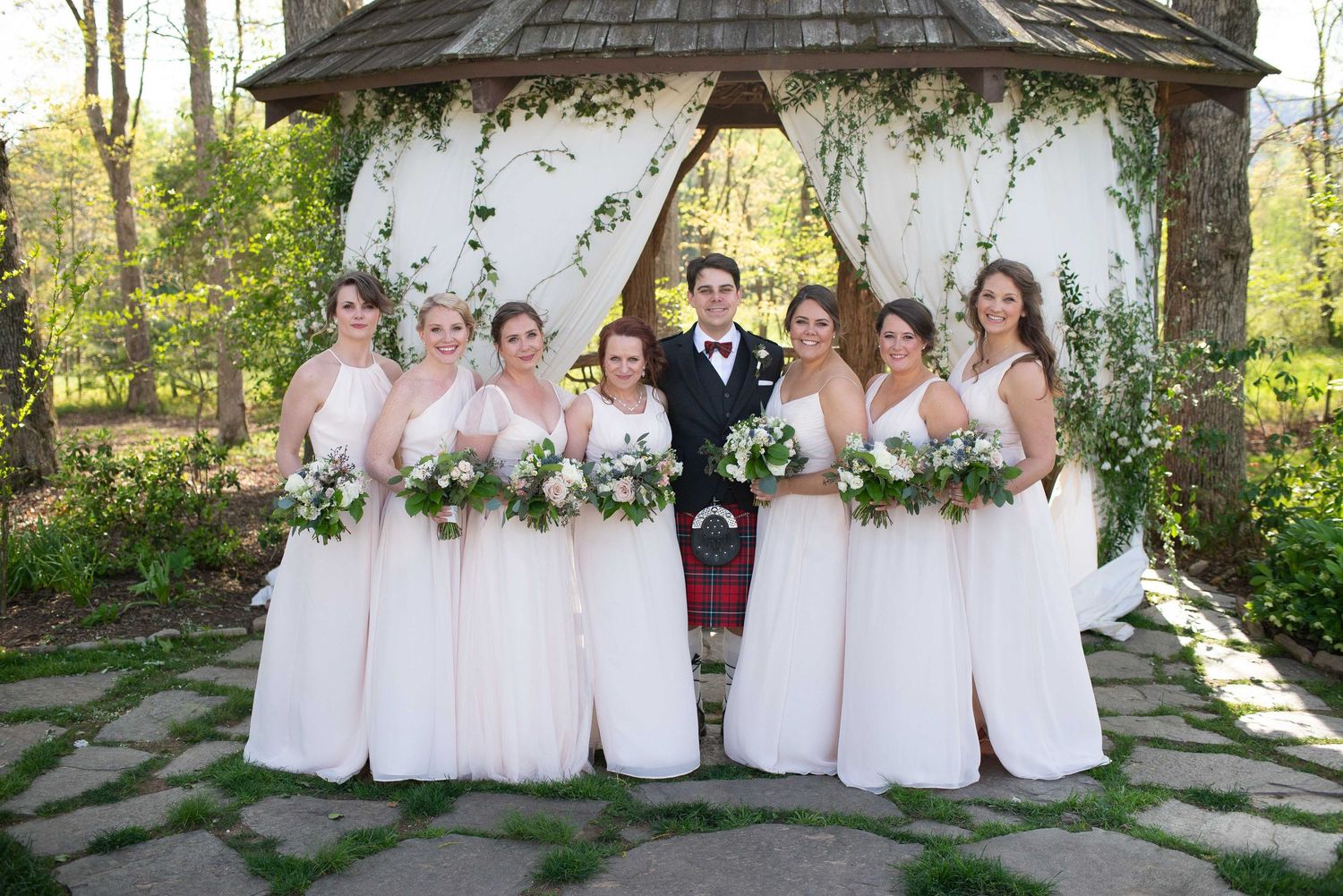 Wedding party poses under floral gazebo, bridesmaids in blush gowns flank bride and groom in kilt.