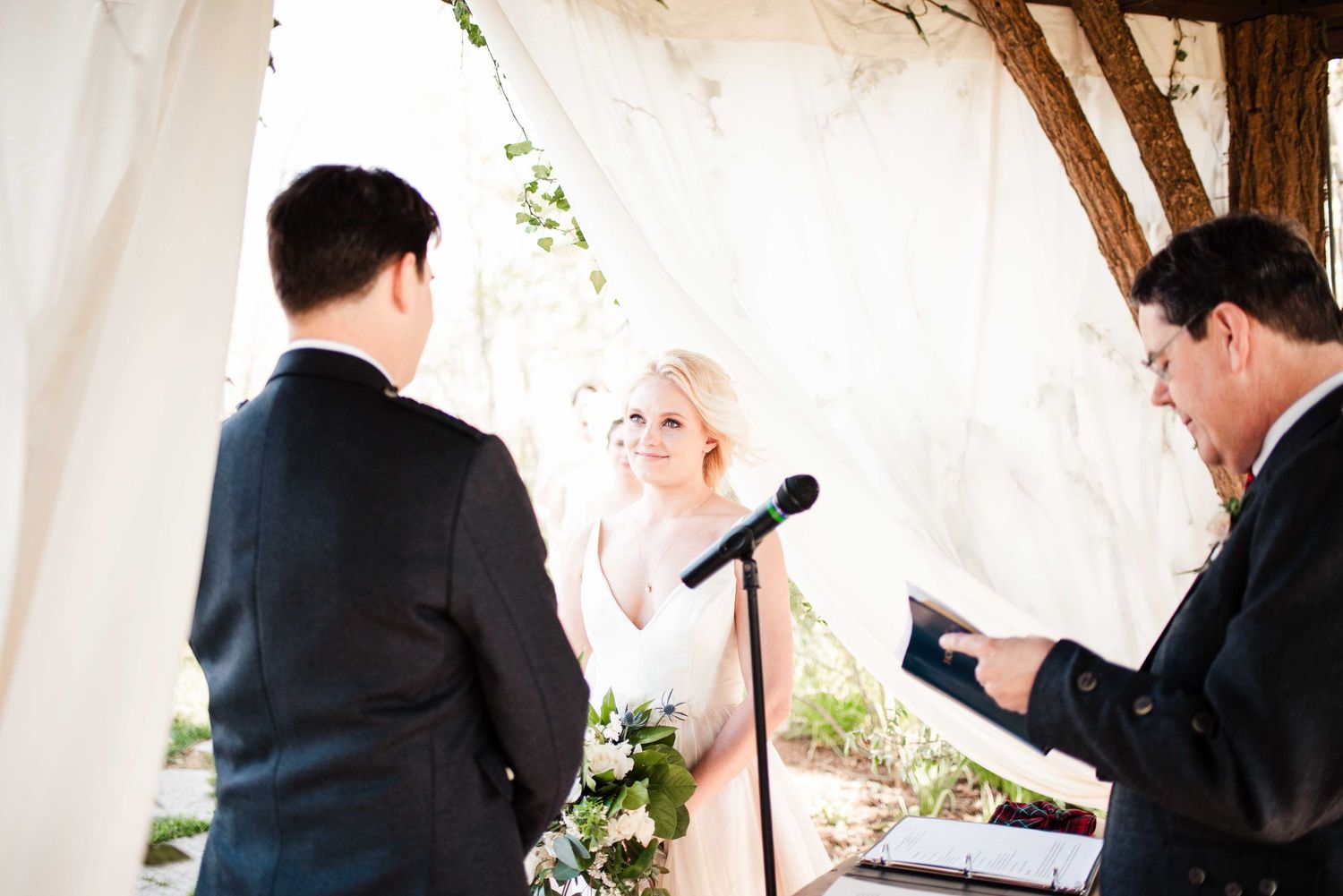 Bride and groom exchange vows at outdoor wedding ceremony with officiant reading from book under white draped canopy.