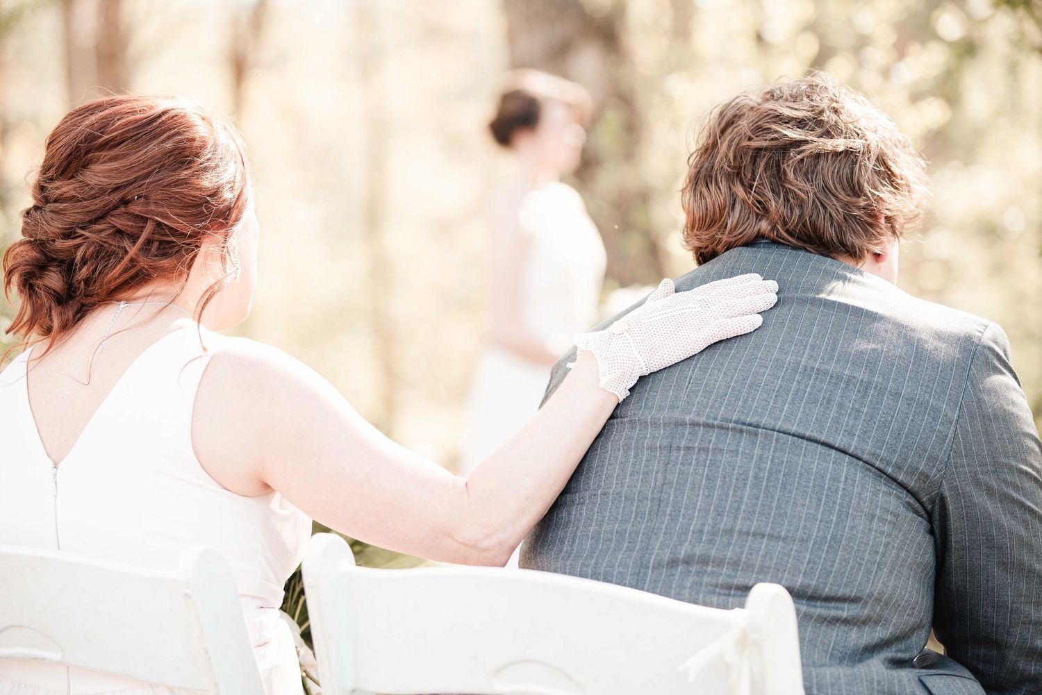 A bride and groom sit together at an outdoor wedding ceremony, viewed from behind in soft natural light.