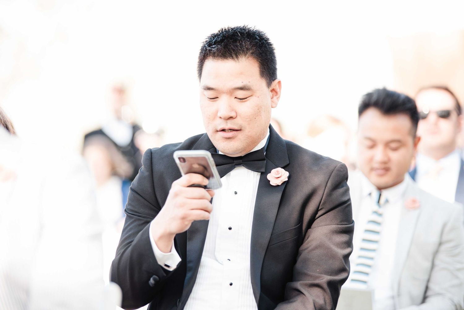 Groom in tuxedo with bow tie and boutonniere checking phone at outdoor wedding ceremony.