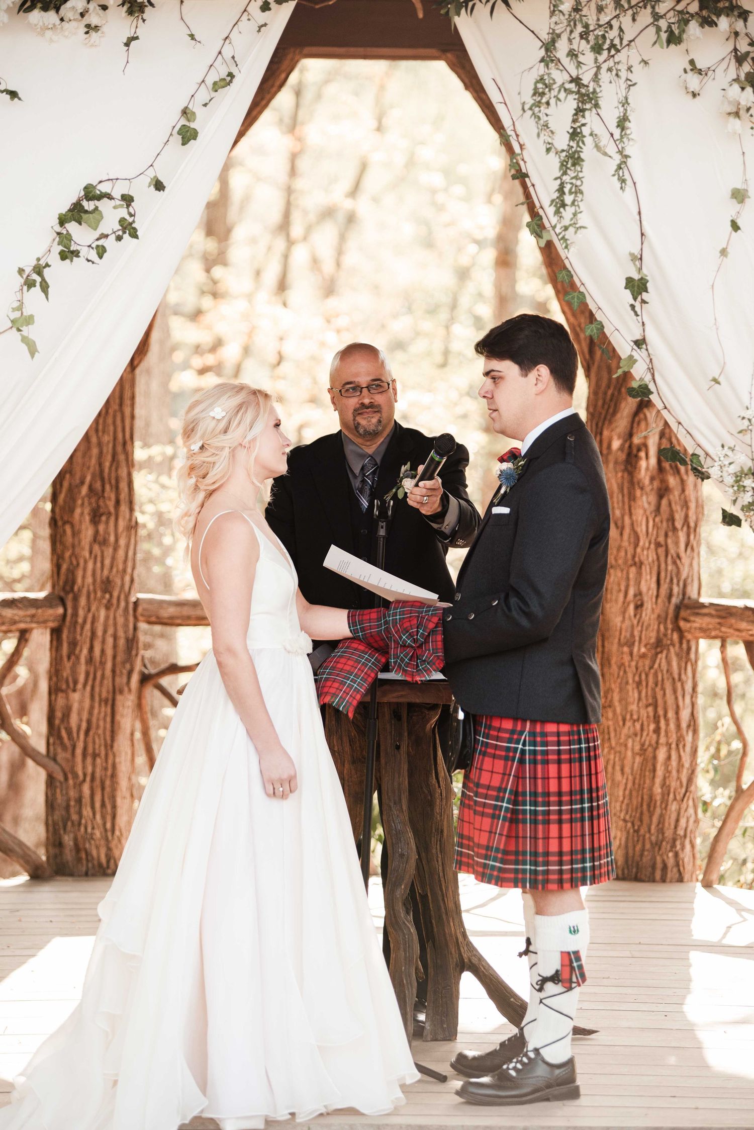 Outdoor wedding ceremony with bride in white gown and groom in red tartan kilt standing before officiant under wooden arch.