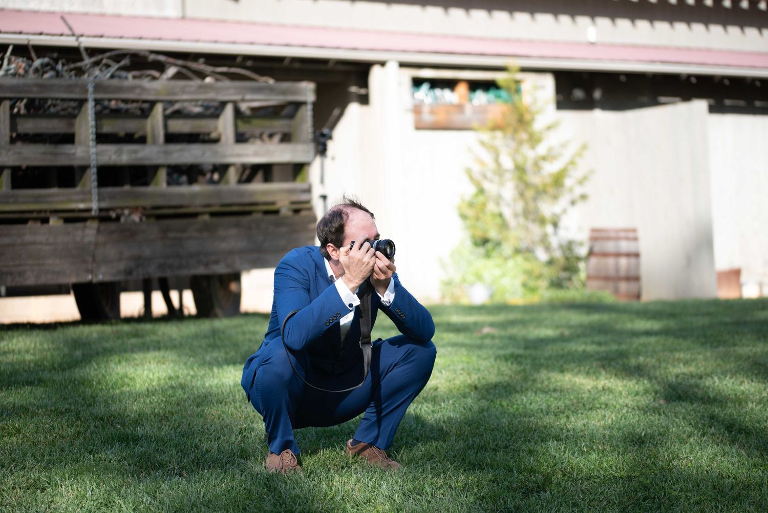 Man in blue suit crouching on grass, taking a photo with a camera outdoors near a wooden structure.