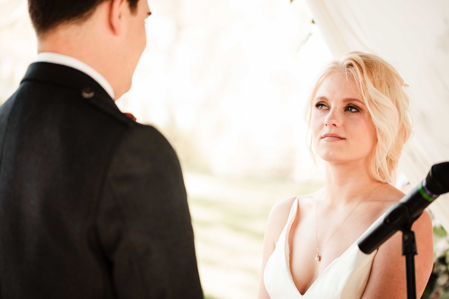 Bride gazing lovingly at groom during outdoor wedding ceremony, wearing white dress near microphone stand.