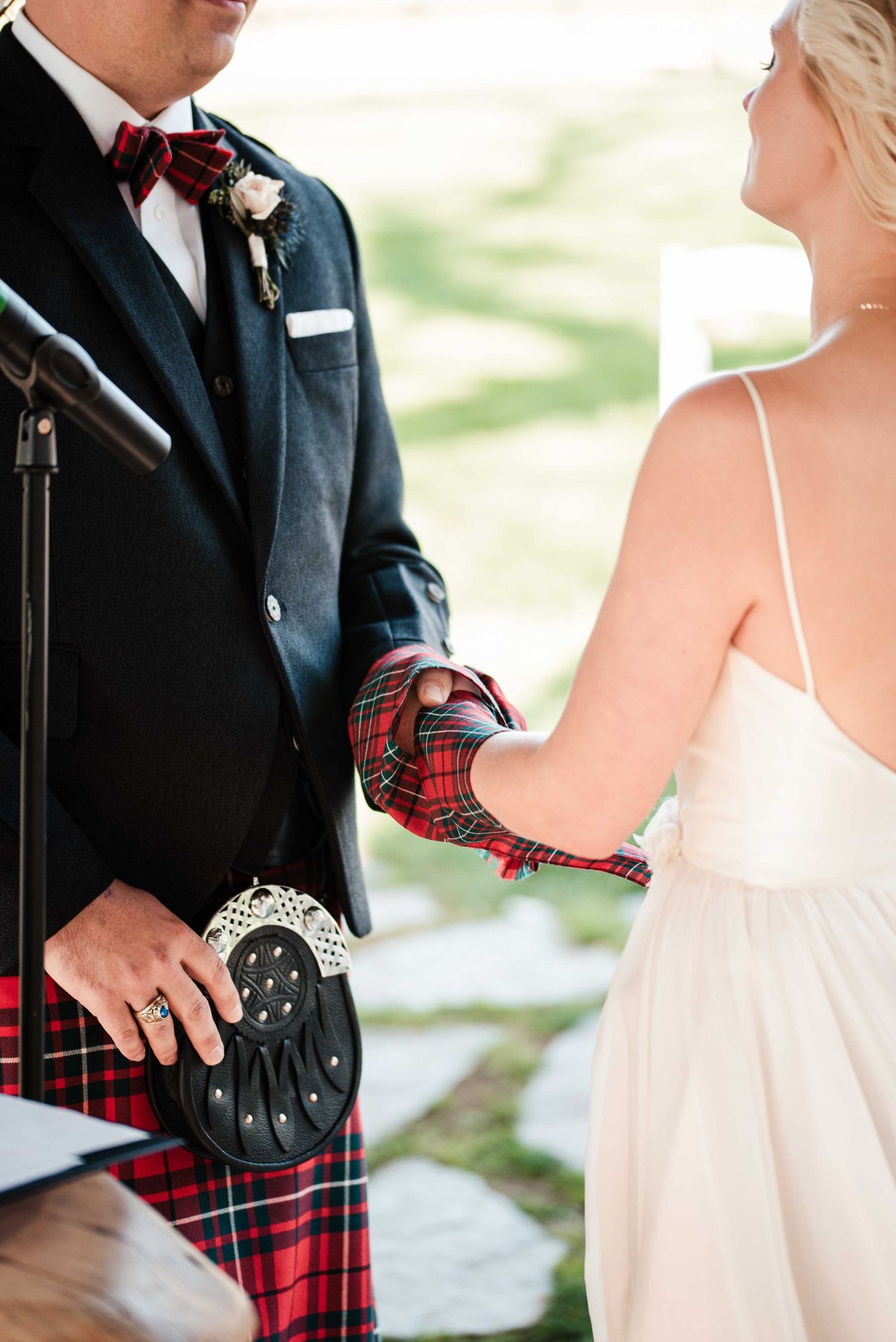 Groom in red tartan kilt and navy blazer holds hands with bride in white dress at outdoor wedding ceremony.