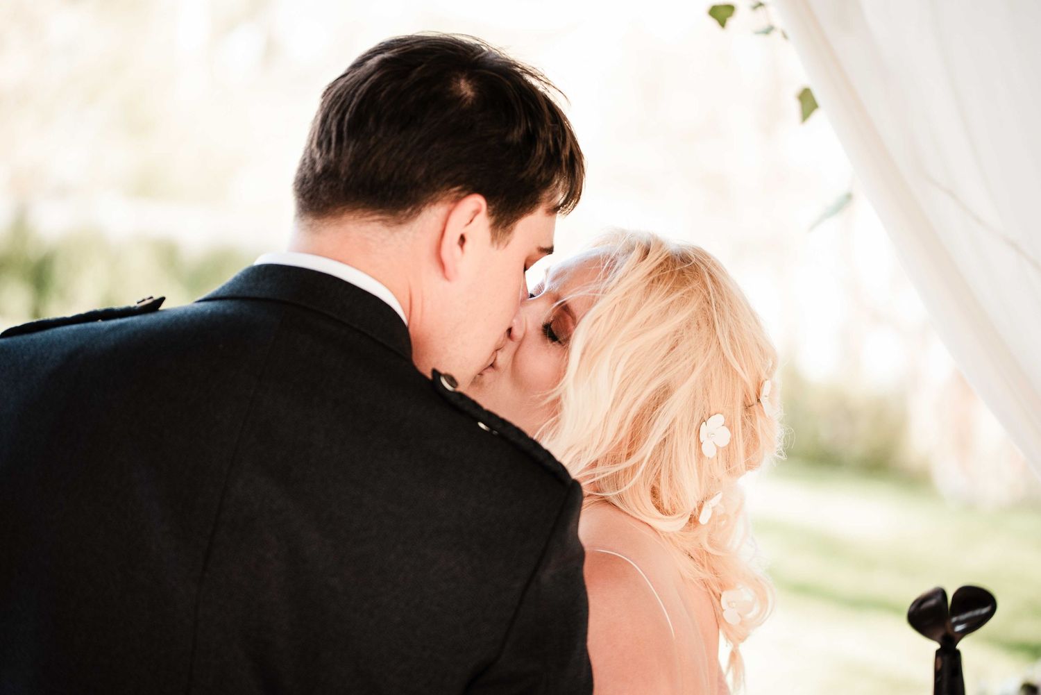 A bride and groom share their first kiss at an outdoor wedding ceremony beneath a white draped arch.