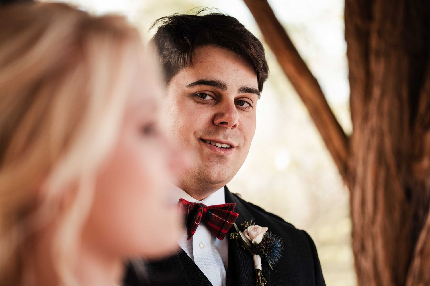 Groom in black suit and red plaid bow tie smiling warmly at blurred blonde bride during wedding ceremony.