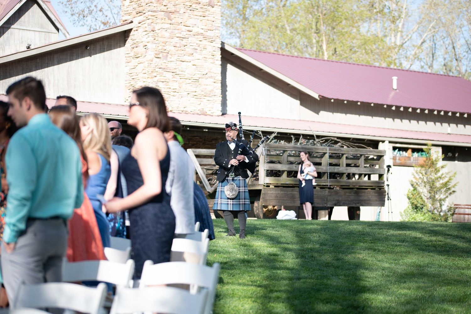 A bagpiper performs outdoors at a rustic wedding venue while guests in formal attire stand on a green lawn.