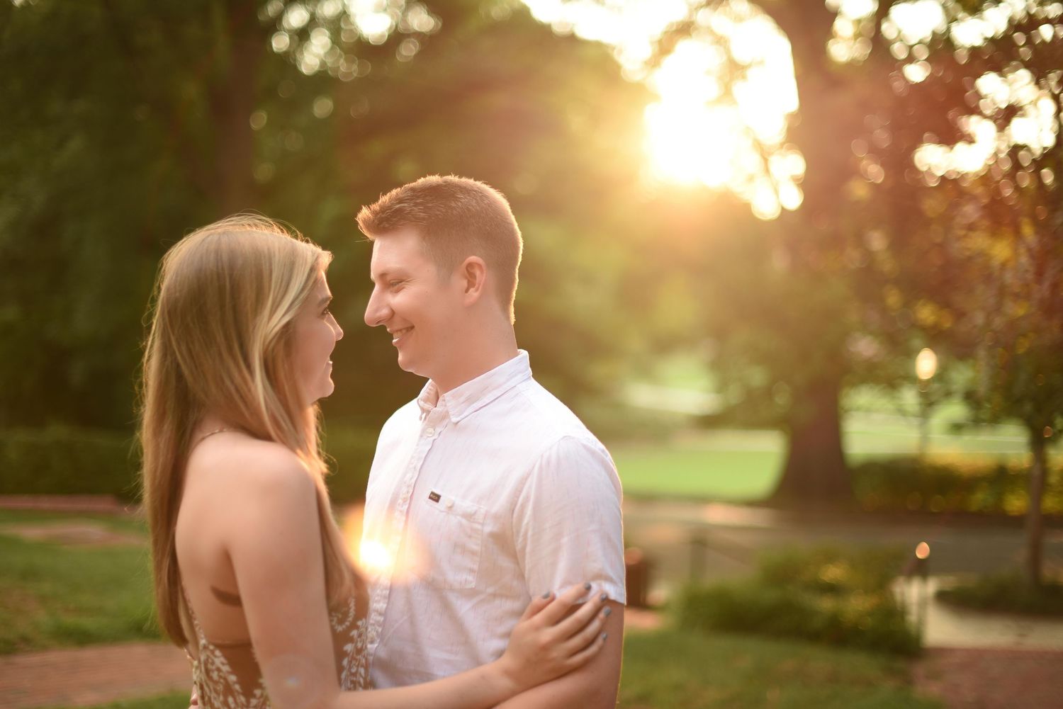 Couple shares a tender moment at golden hour with warm sunlight glowing through trees in a park.