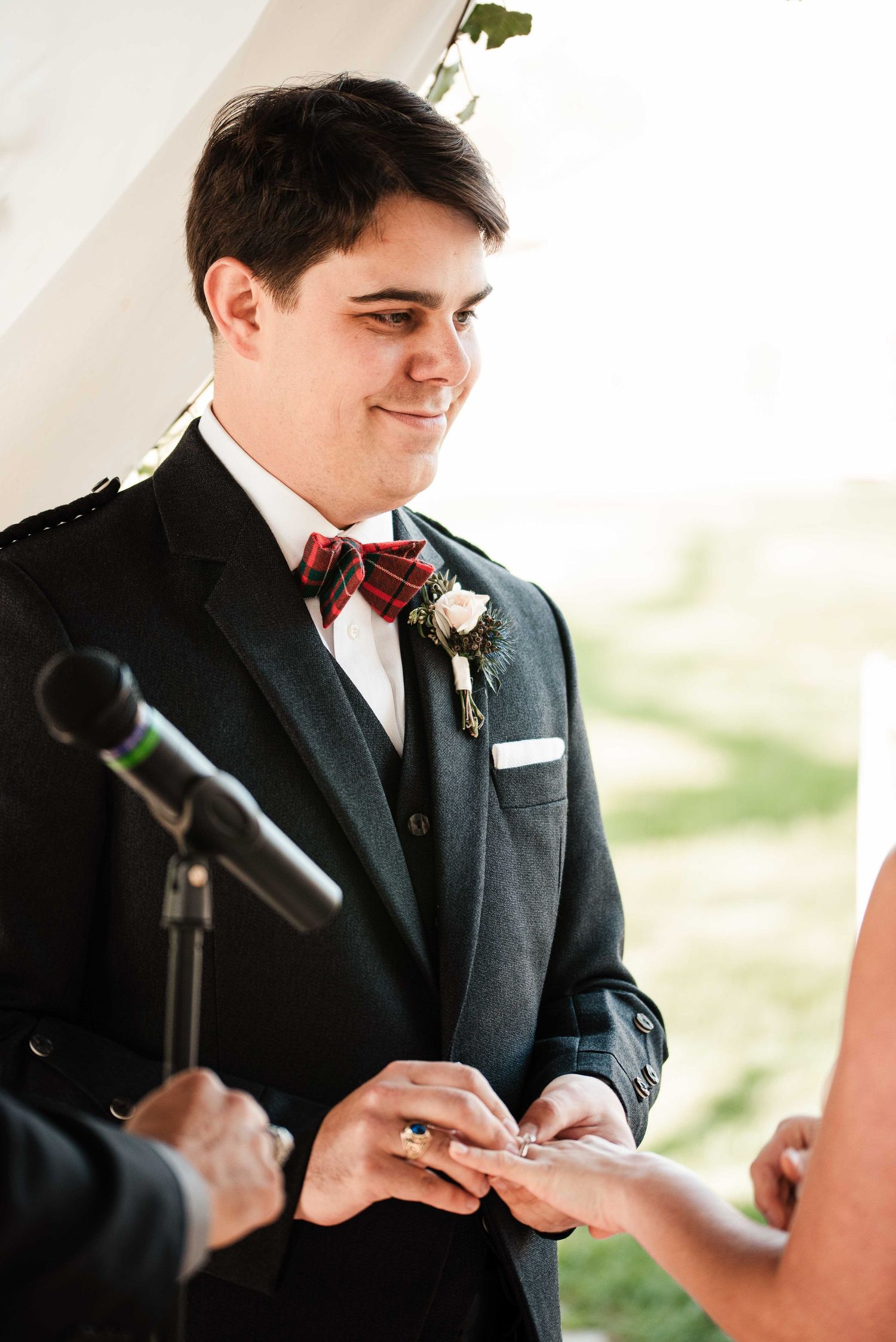 Smiling groom in plaid bow tie exchanges rings during outdoor wedding ceremony under tent.