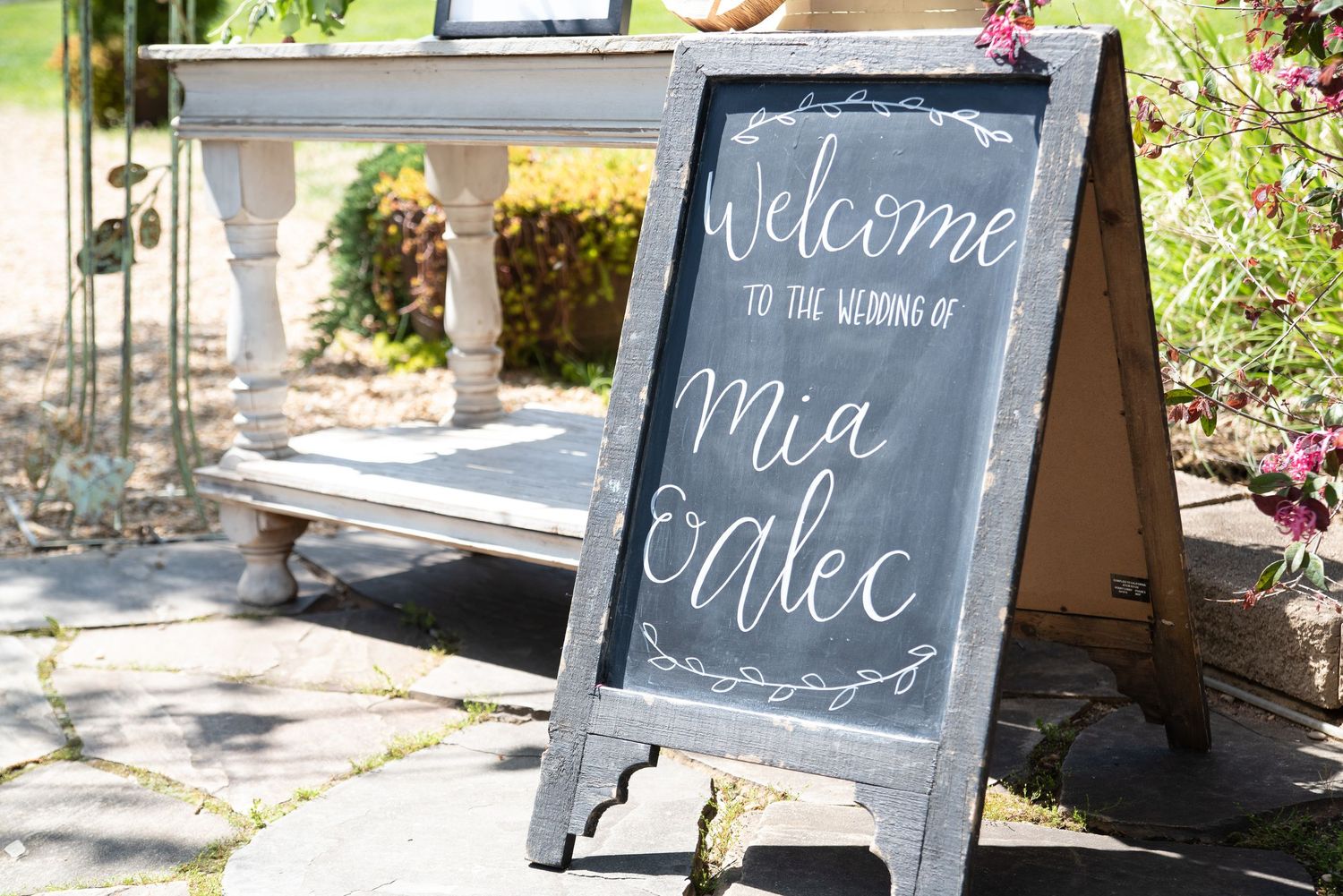 A chalkboard welcome sign reads Welcome to the Wedding of Mia and Alec at an outdoor wedding venue.