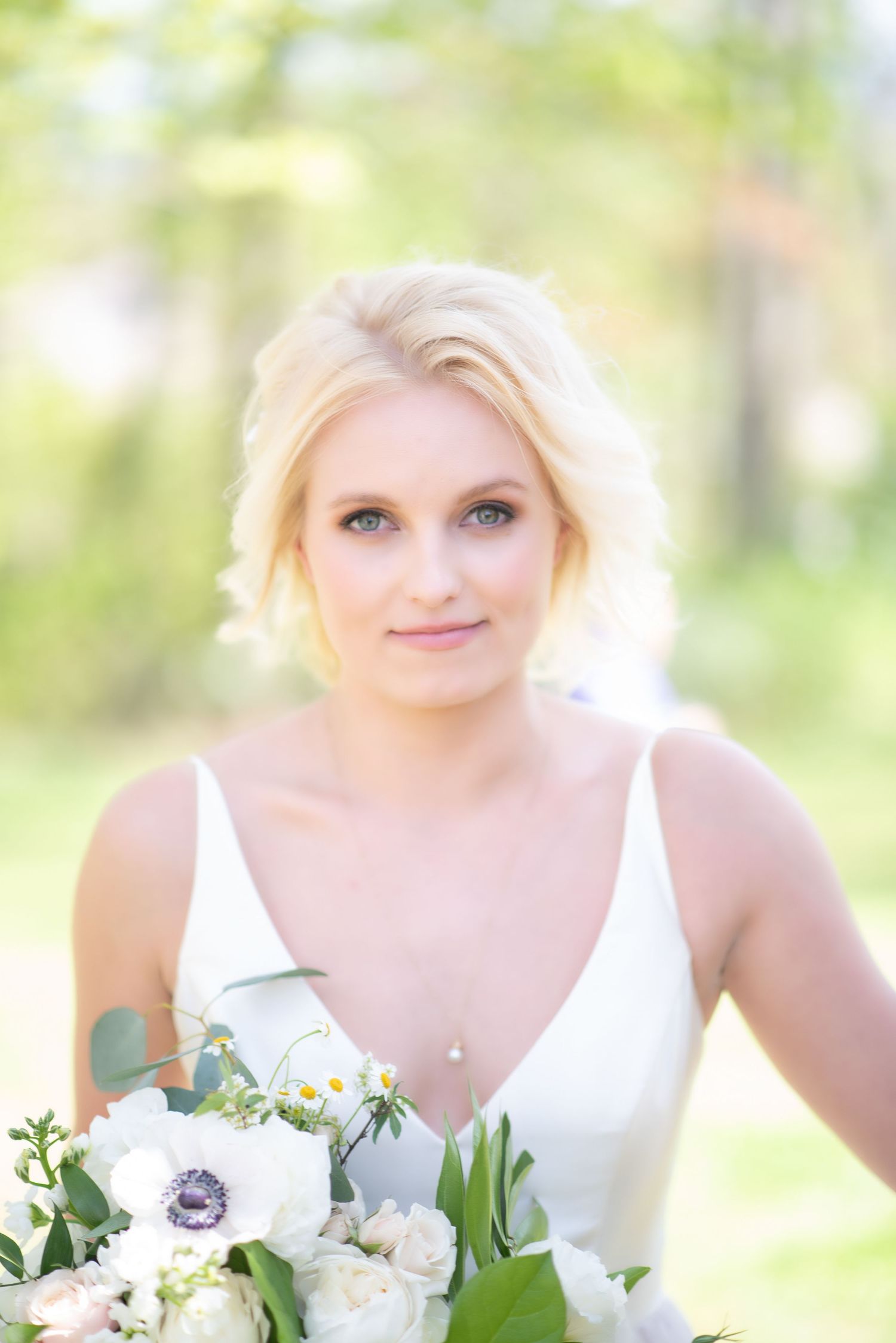 Beautiful blonde bride in white gown holding a white floral bouquet outdoors in soft natural light.