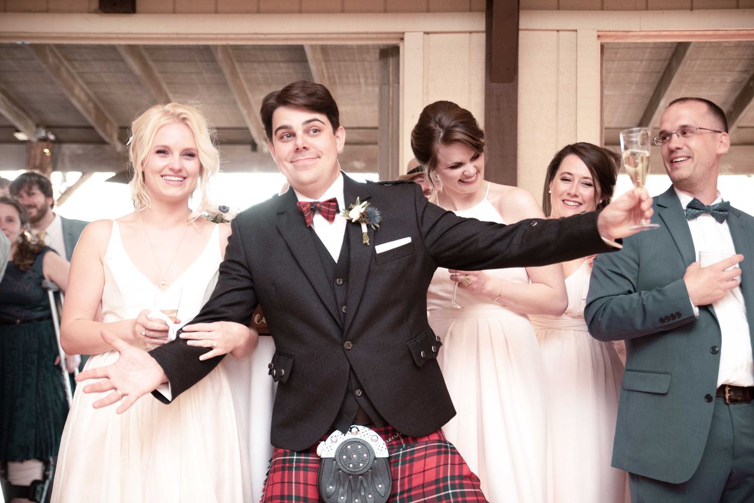 Groom in Scottish kilt and bow tie celebrates with bridesmaids in white dresses at wedding reception.