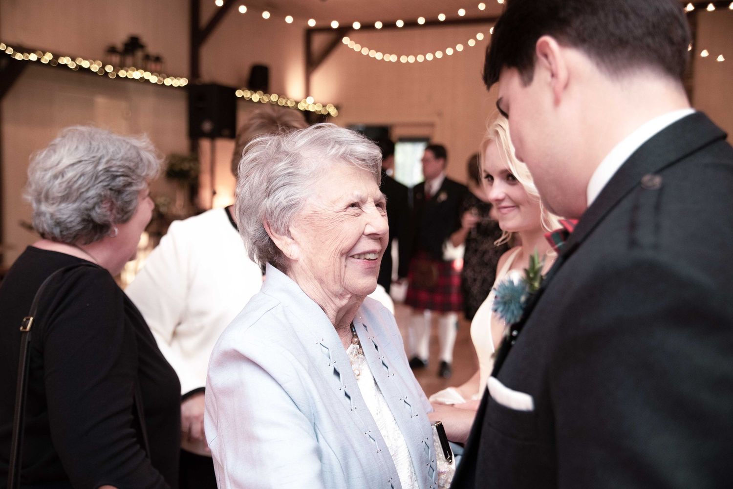 Elderly woman in blue jacket smiles warmly at wedding reception with string lights in background.