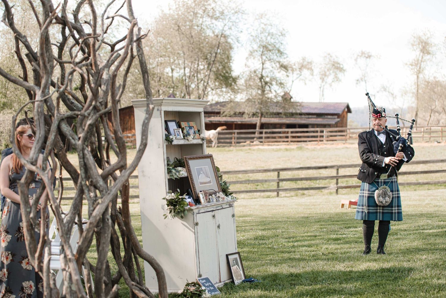 Bagpiper in tartan kilt performs outdoors at rustic farm wedding venue beside decorative branch display.