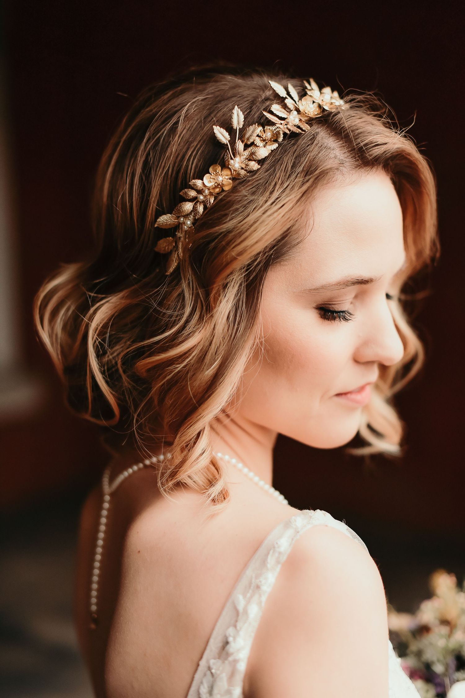 Bride with wavy updo adorned with a delicate gold leaf crown and pearl necklace gazes downward softly.
