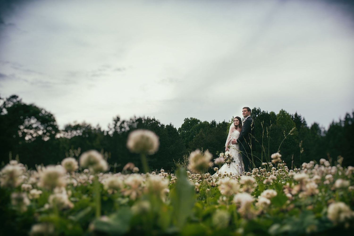 Romantic couple standing in white wildflower field under dramatic cloudy sky at wedding photo shoot.