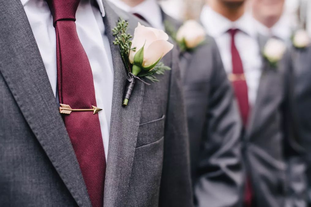 Groomsmen in gray suits with burgundy ties and rose boutonnieres with gold arrow tie clips at wedding.