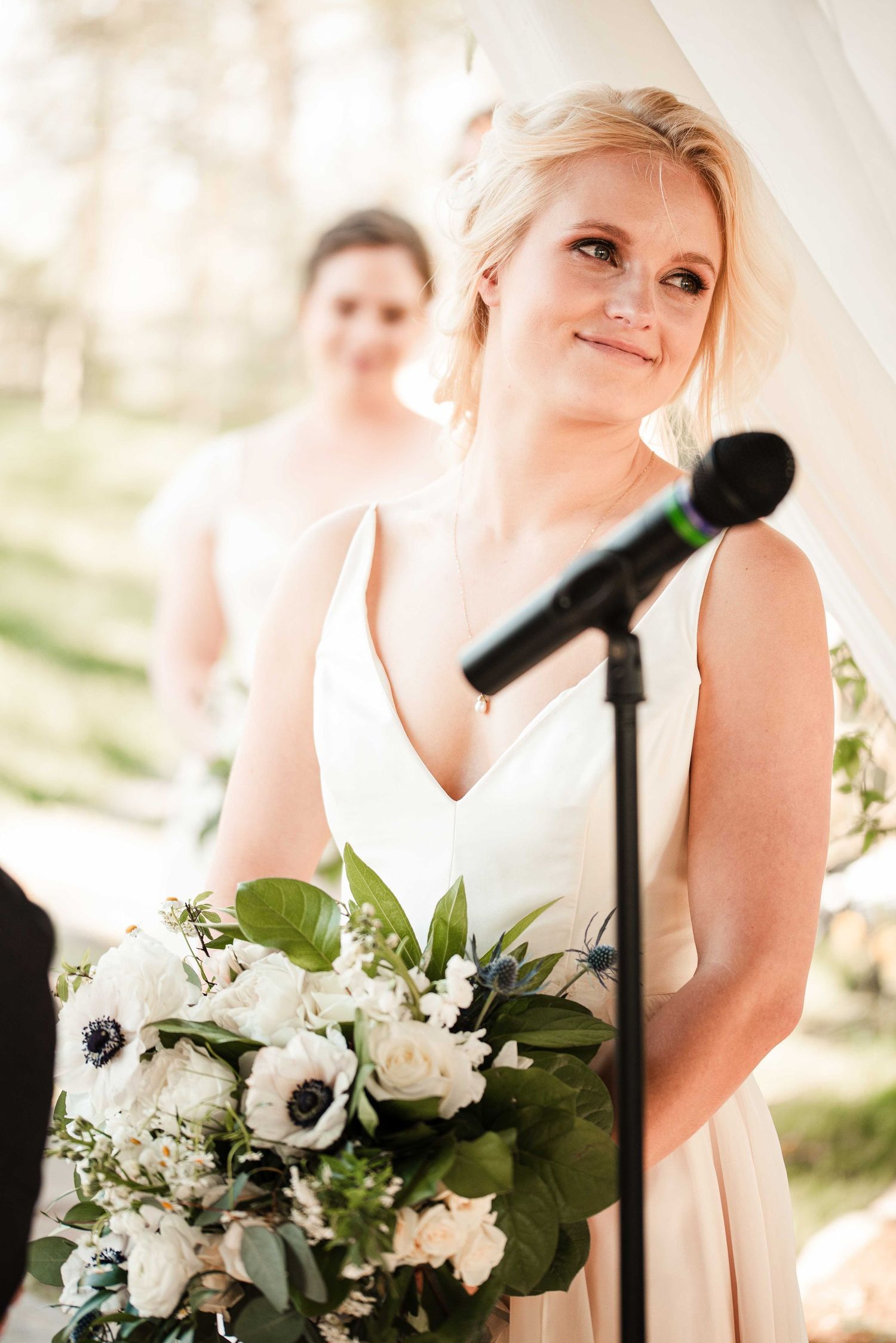 Bride holding white floral bouquet with greenery smiles at wedding ceremony near microphone stand.