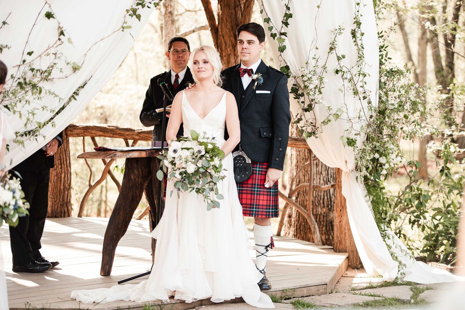 Bride in white gown and groom in red tartan kilt stand at outdoor wedding altar with floral arch.