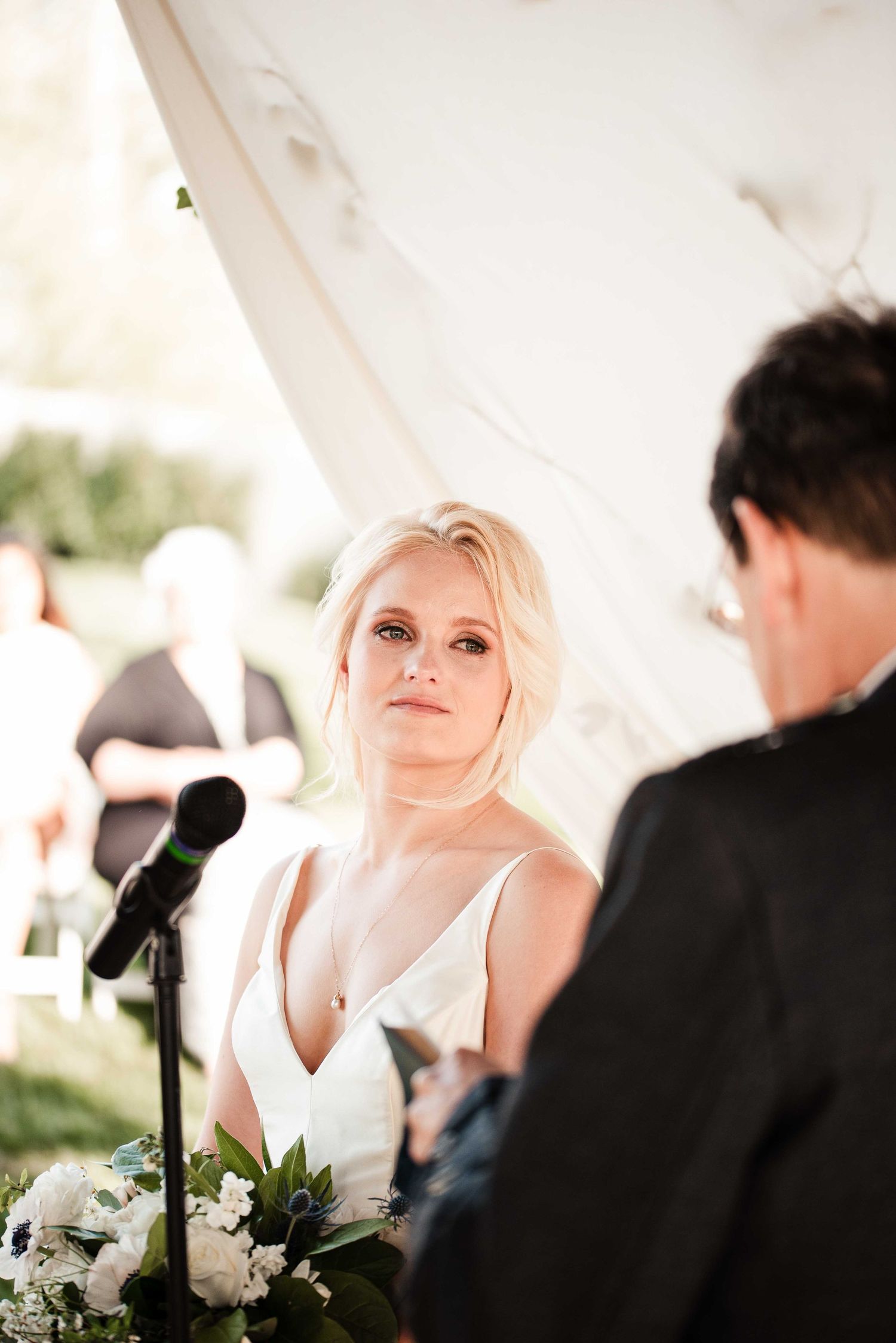 Bride gazing lovingly at groom during outdoor wedding ceremony under white tent with floral arrangements.