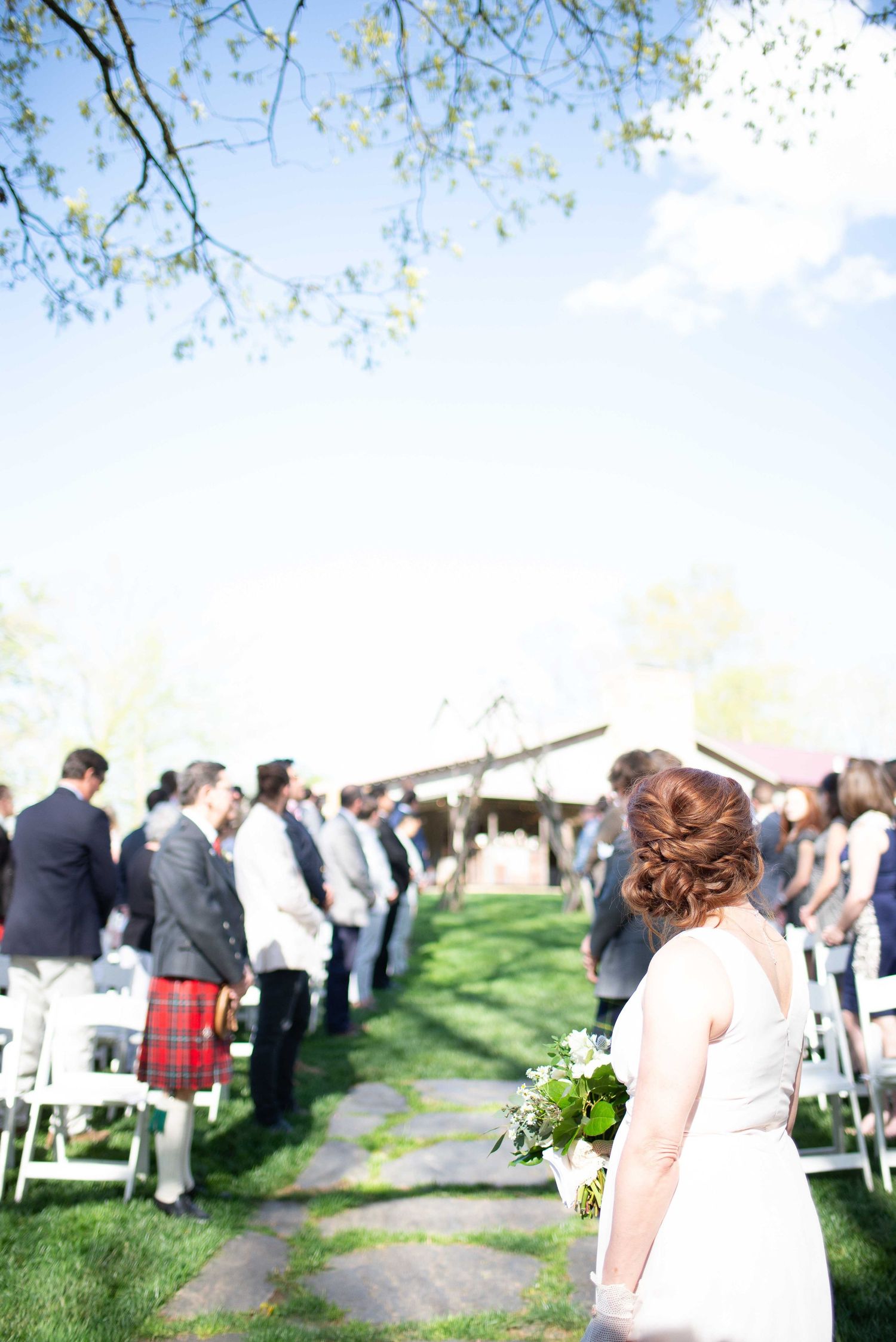 Outdoor wedding ceremony on a sunny day with guests standing along a stone aisle, bride holding white bouquet in foreground.