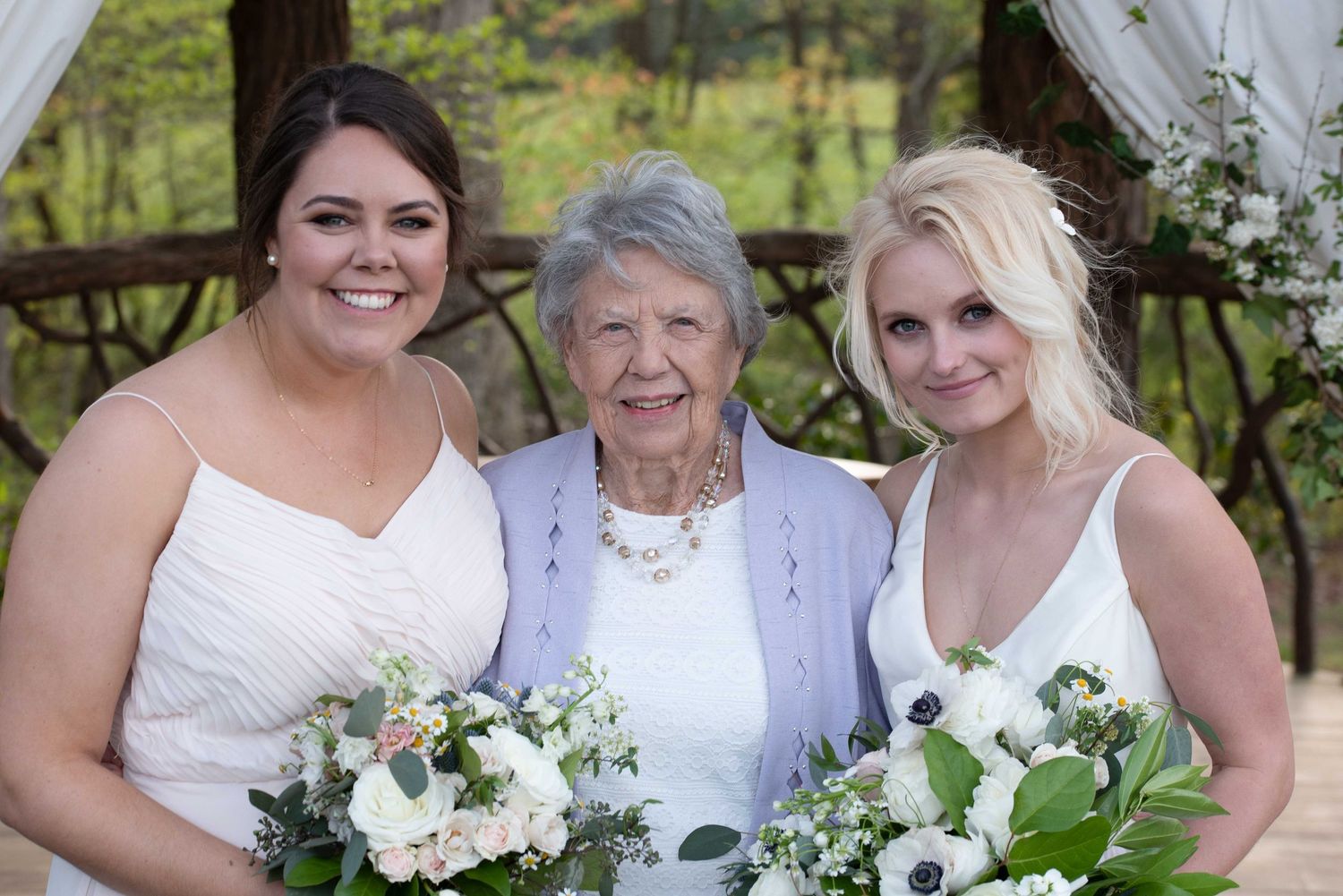 Two bridesmaids in white dresses holding floral bouquets pose with an elderly woman at an outdoor wedding ceremony.