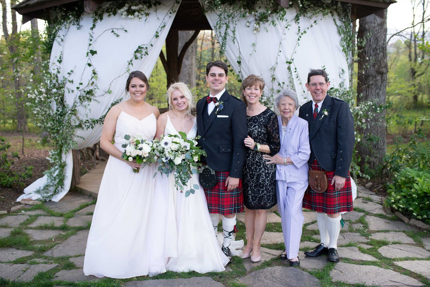 Wedding party poses under floral arch, bride in white gown, groom in kilt, family in formal attire outdoors.