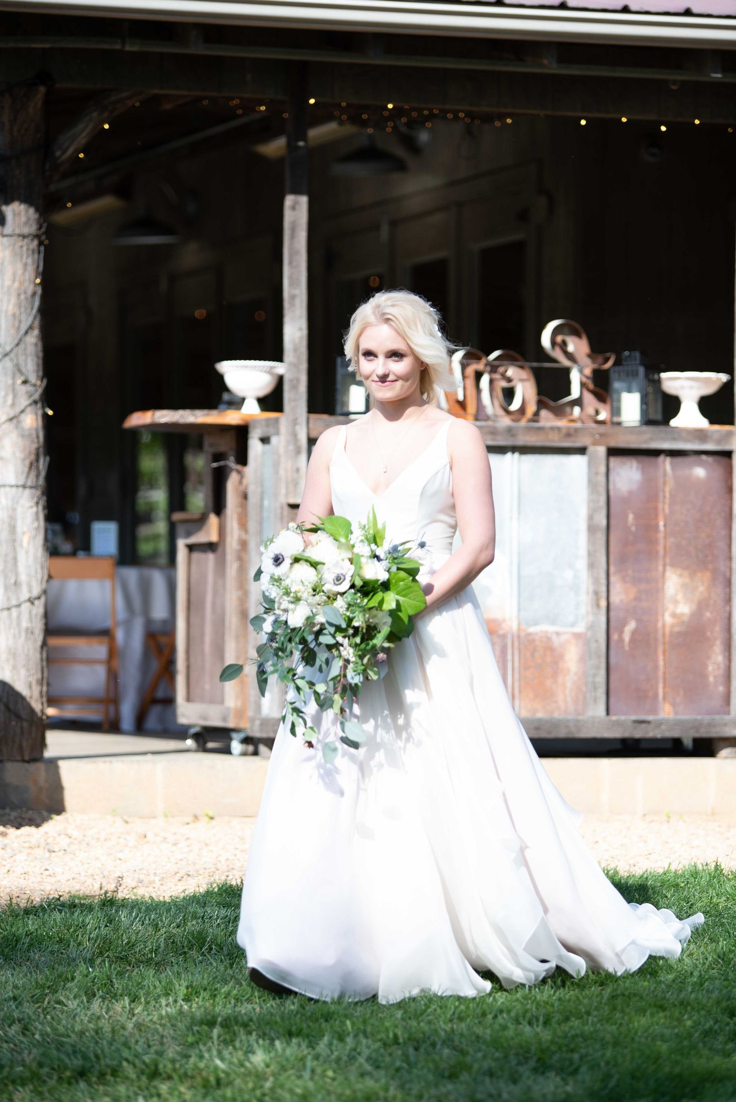 Bride in white A-line gown holding cascading greenery bouquet stands on grass before rustic barn wedding venue.