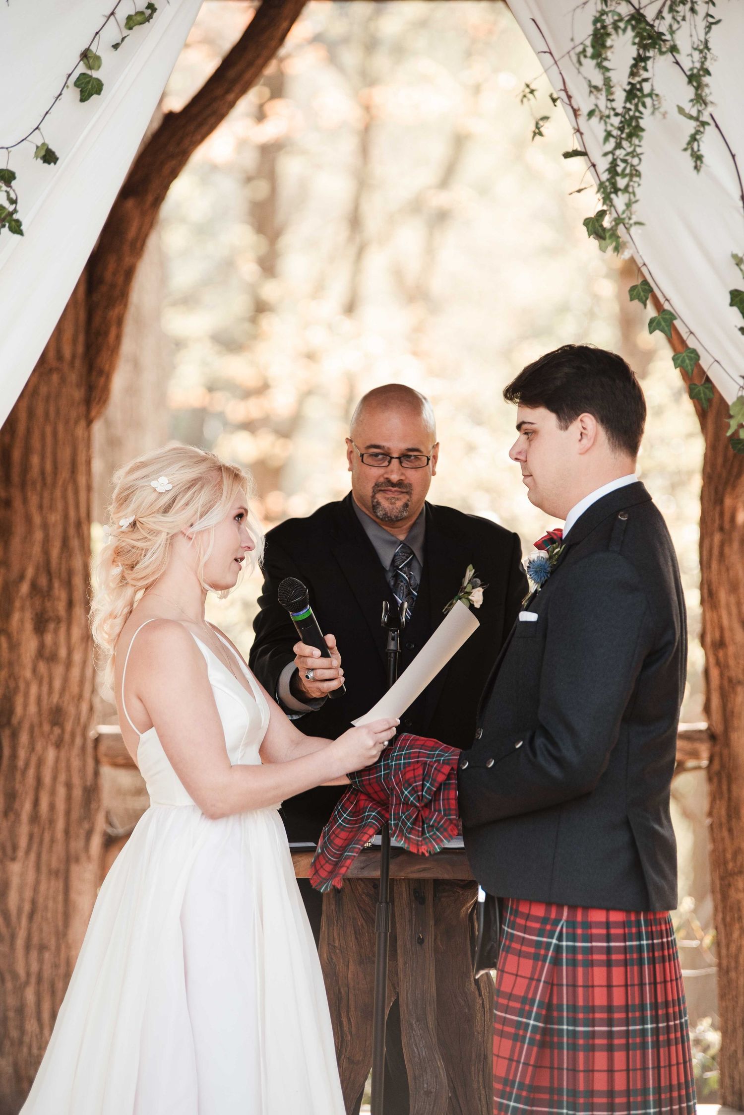 Bride reads vows to groom in red tartan kilt at outdoor wedding ceremony beneath floral arch.