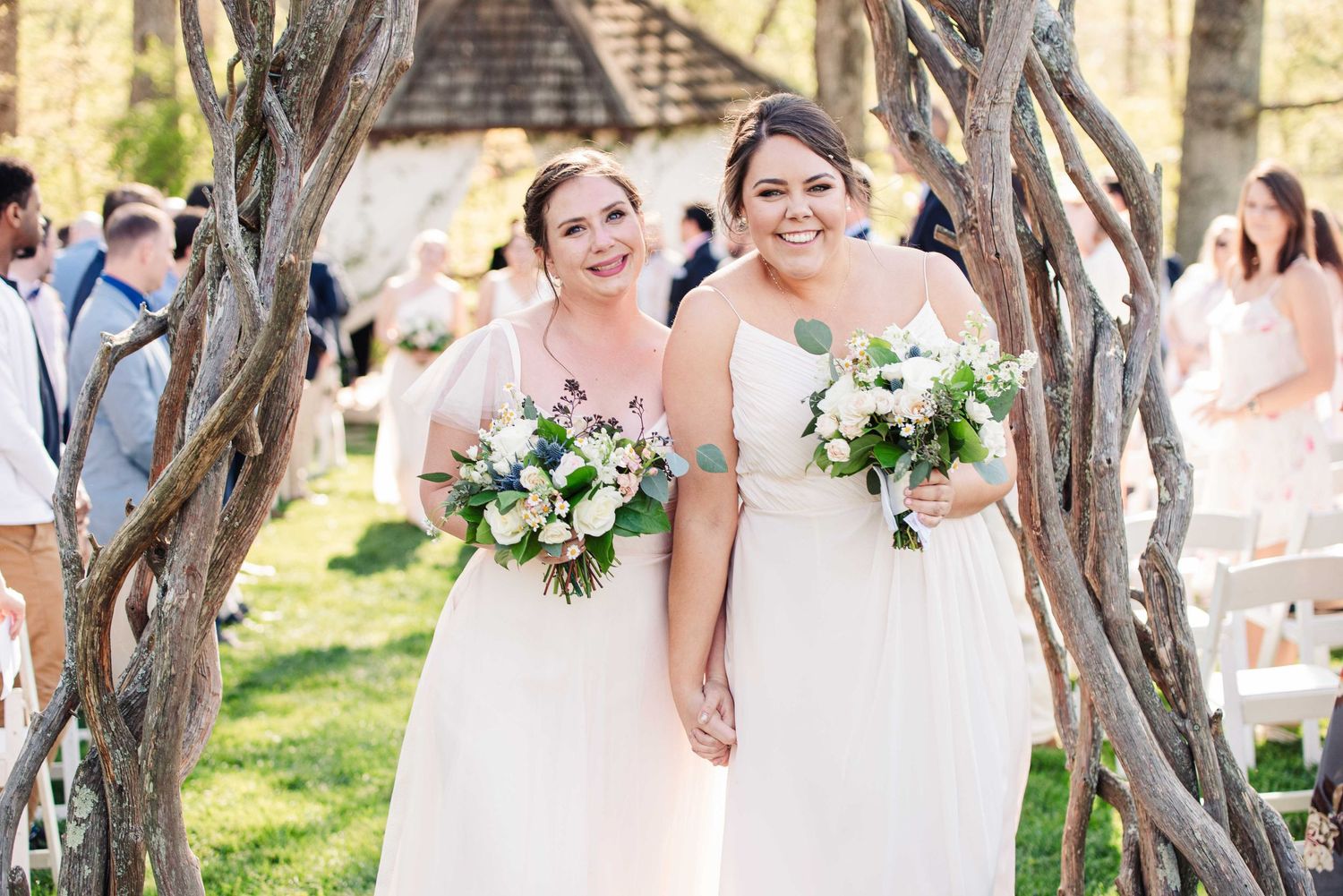 Two brides in white gowns holding bouquets smile under rustic branch arch at outdoor wedding ceremony.