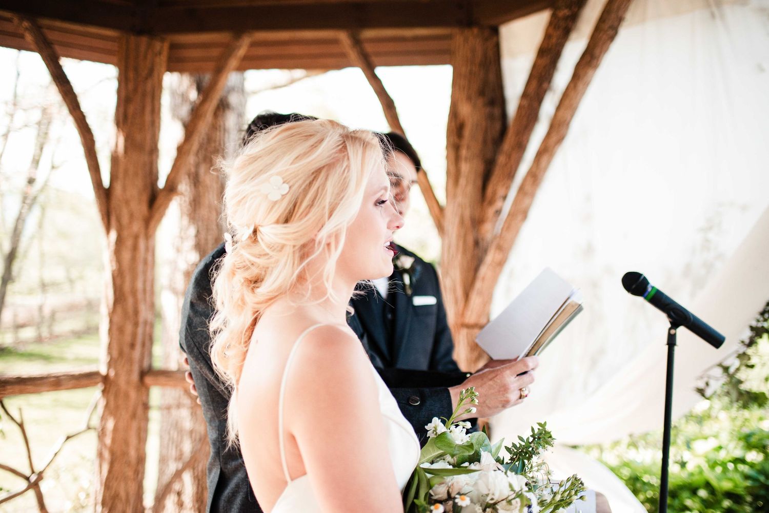 Bride reading vows at outdoor wedding ceremony under rustic wooden gazebo with groom beside her.