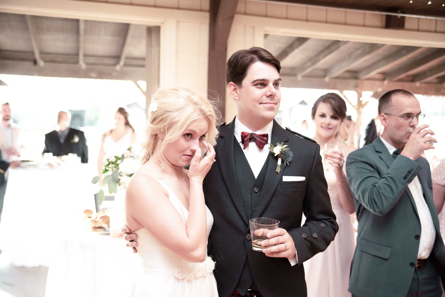 Emotional bride wiping tears while groom smiles during wedding reception toast.