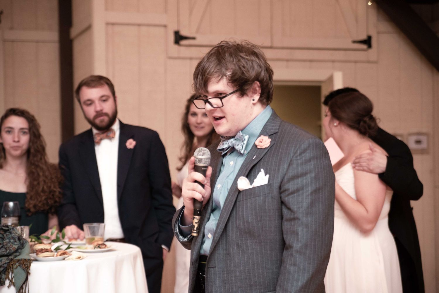 Man in gray suit delivers heartfelt wedding toast with microphone at indoor reception venue.