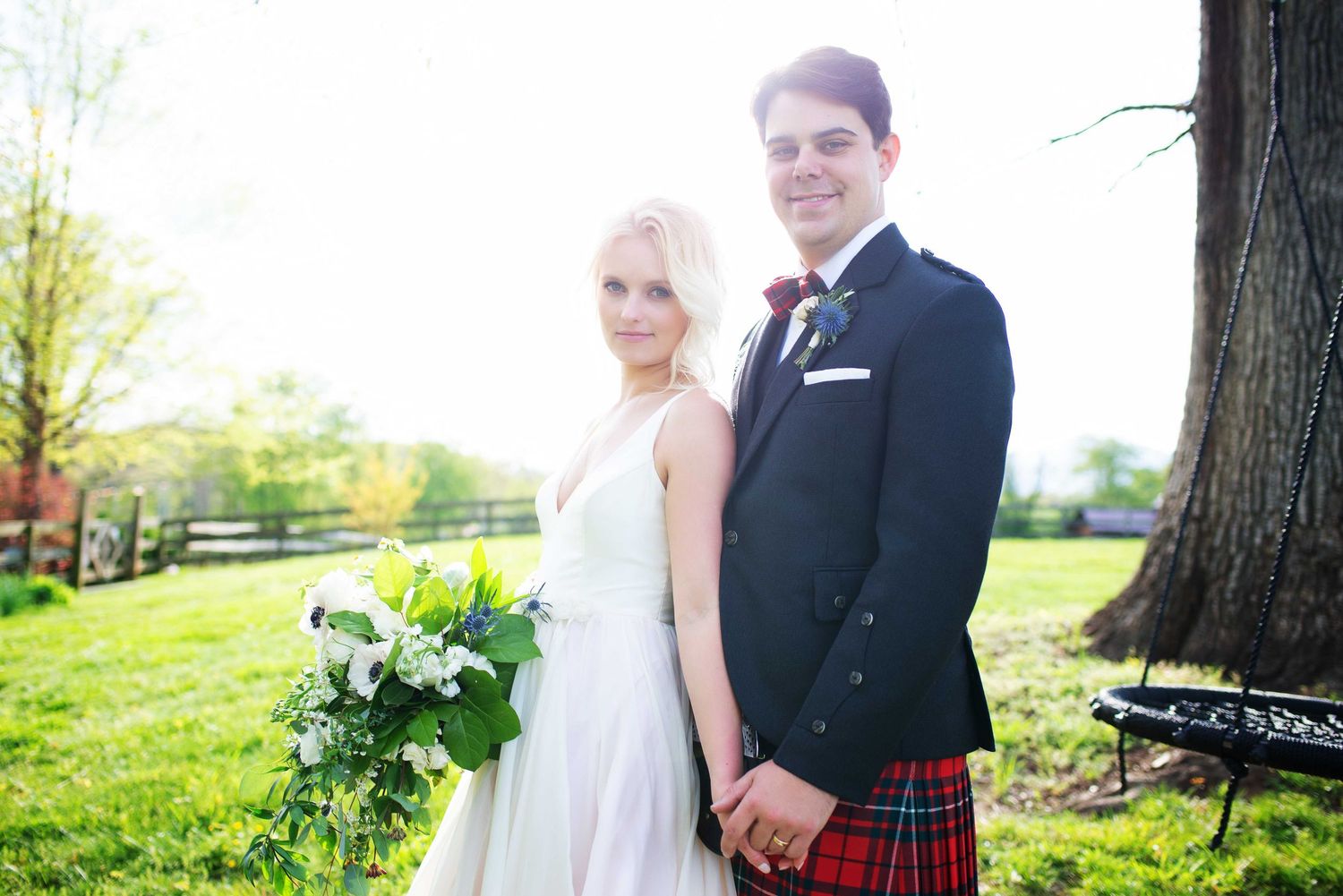 Bride and groom holding hands outdoors, groom wearing red tartan kilt and boutonniere, bride in white gown holding bouquet.