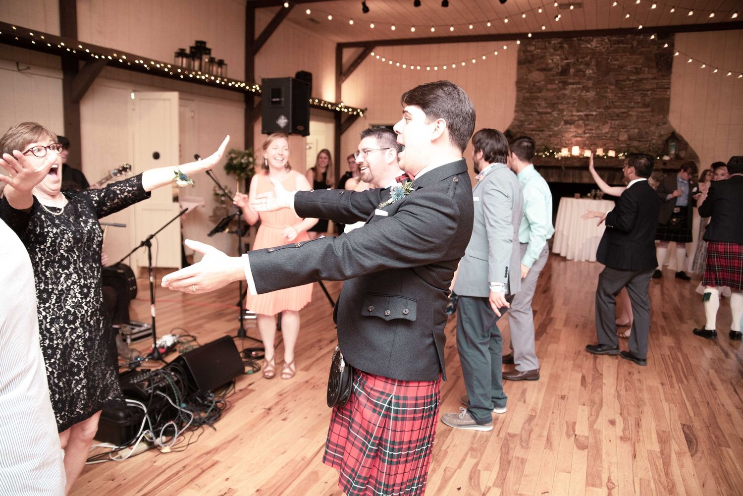 Man in kilt dancing at wedding reception on wooden dance floor with guests and string lights.