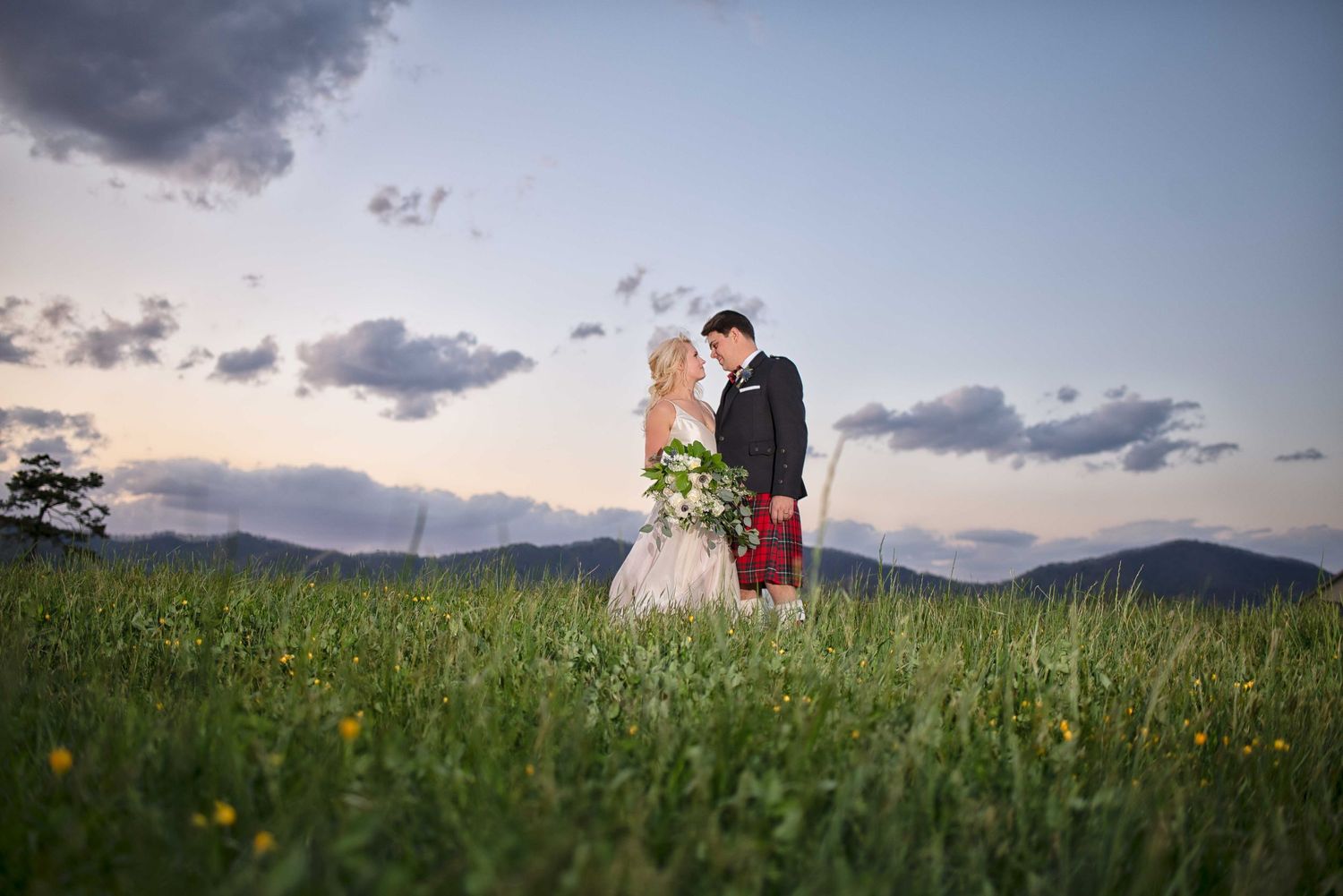 Wedding couple standing in a lush green field at dusk, groom in a Scottish kilt, mountains in the background.