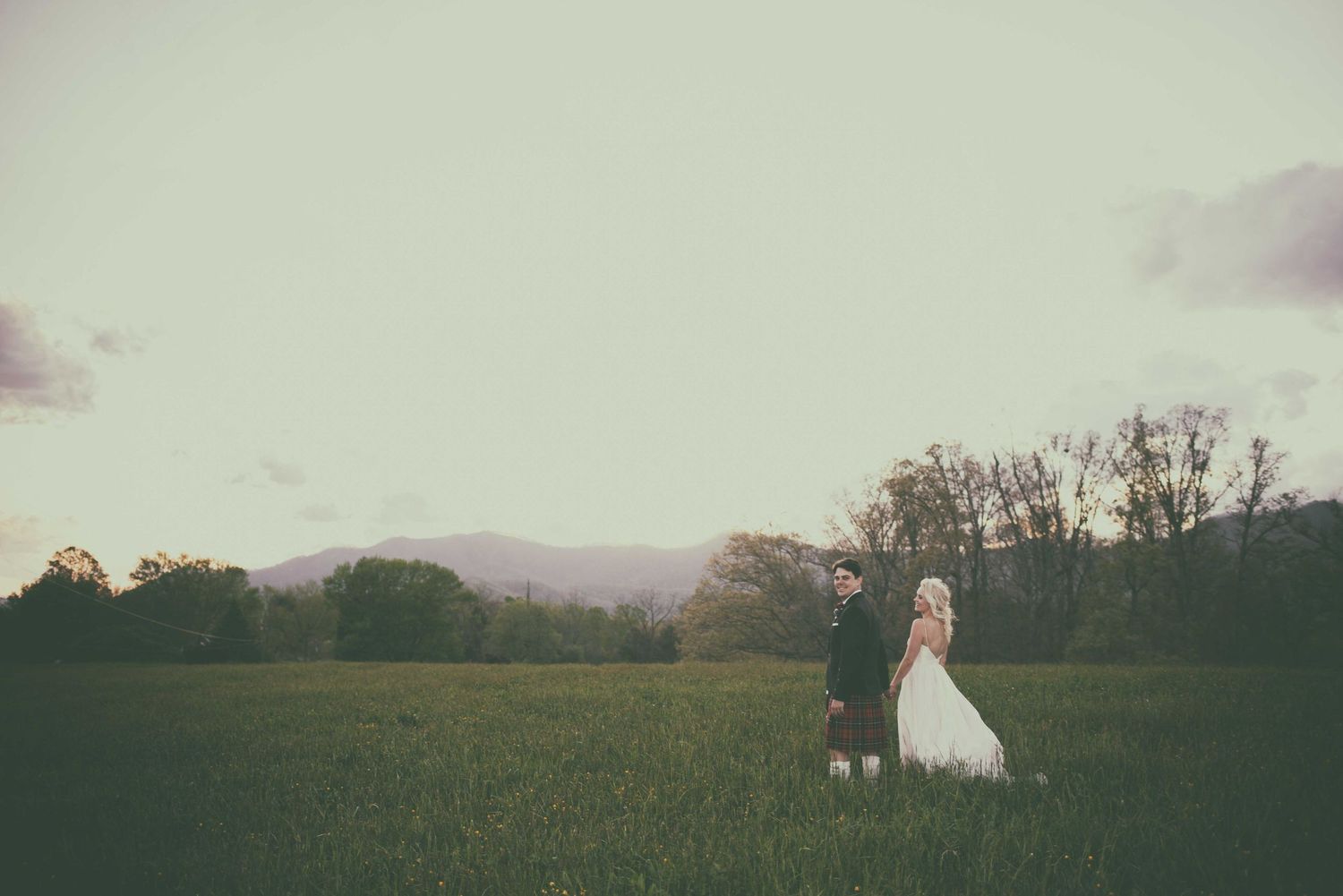 Bride and groom standing in a lush green field with misty mountains and trees in the background.