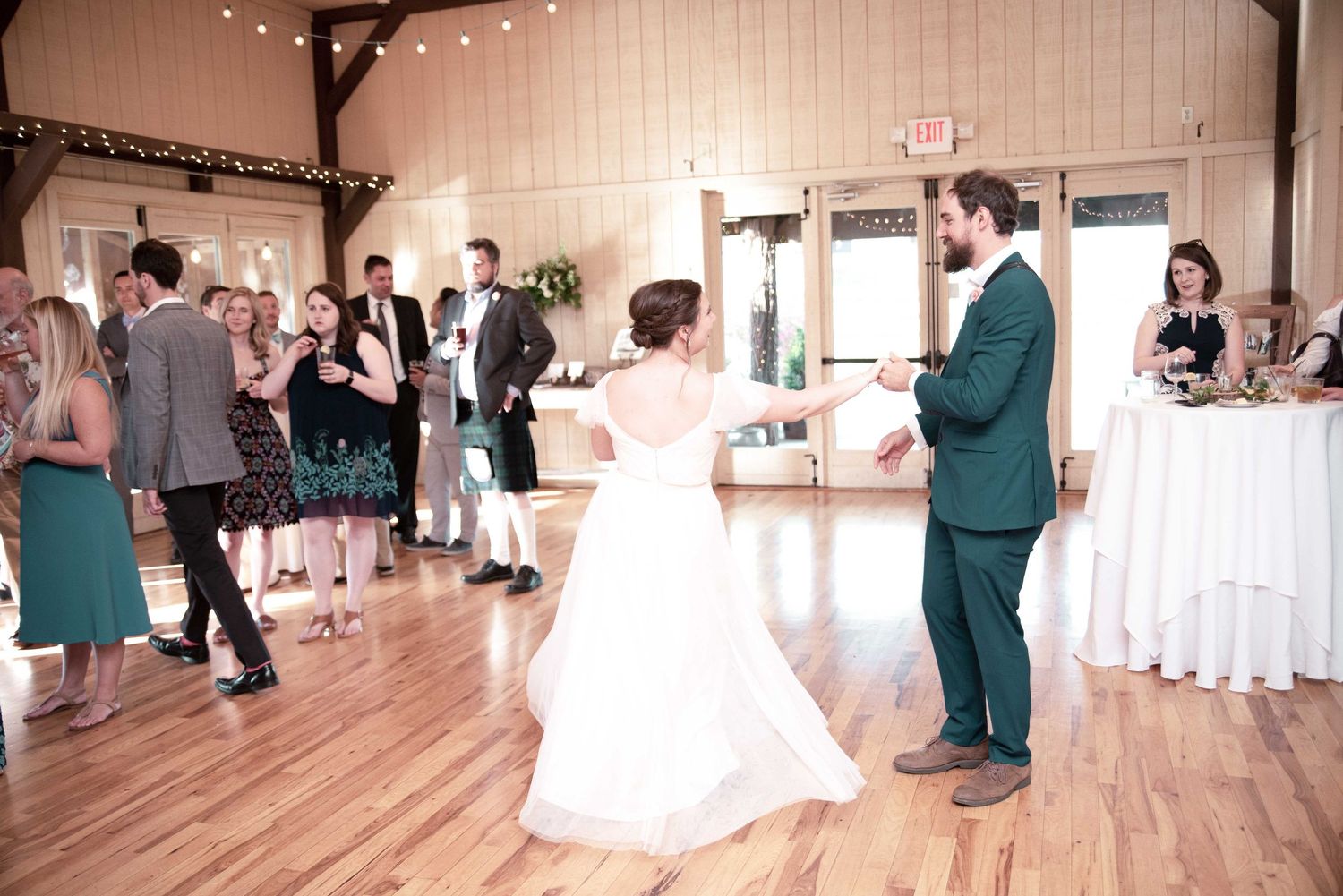 Bride and groom share their first dance at a rustic wedding reception venue with guests watching.