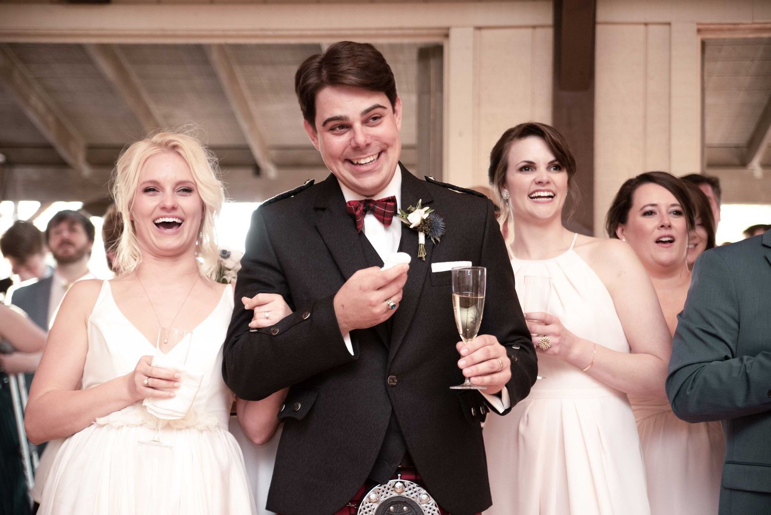 Groom in kilt toasting with champagne flanked by smiling bridesmaids in white dresses at a wedding reception.