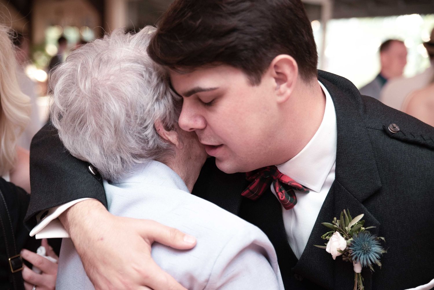 Young man in kilt and bow tie shares tender embrace with elderly woman at a wedding celebration.