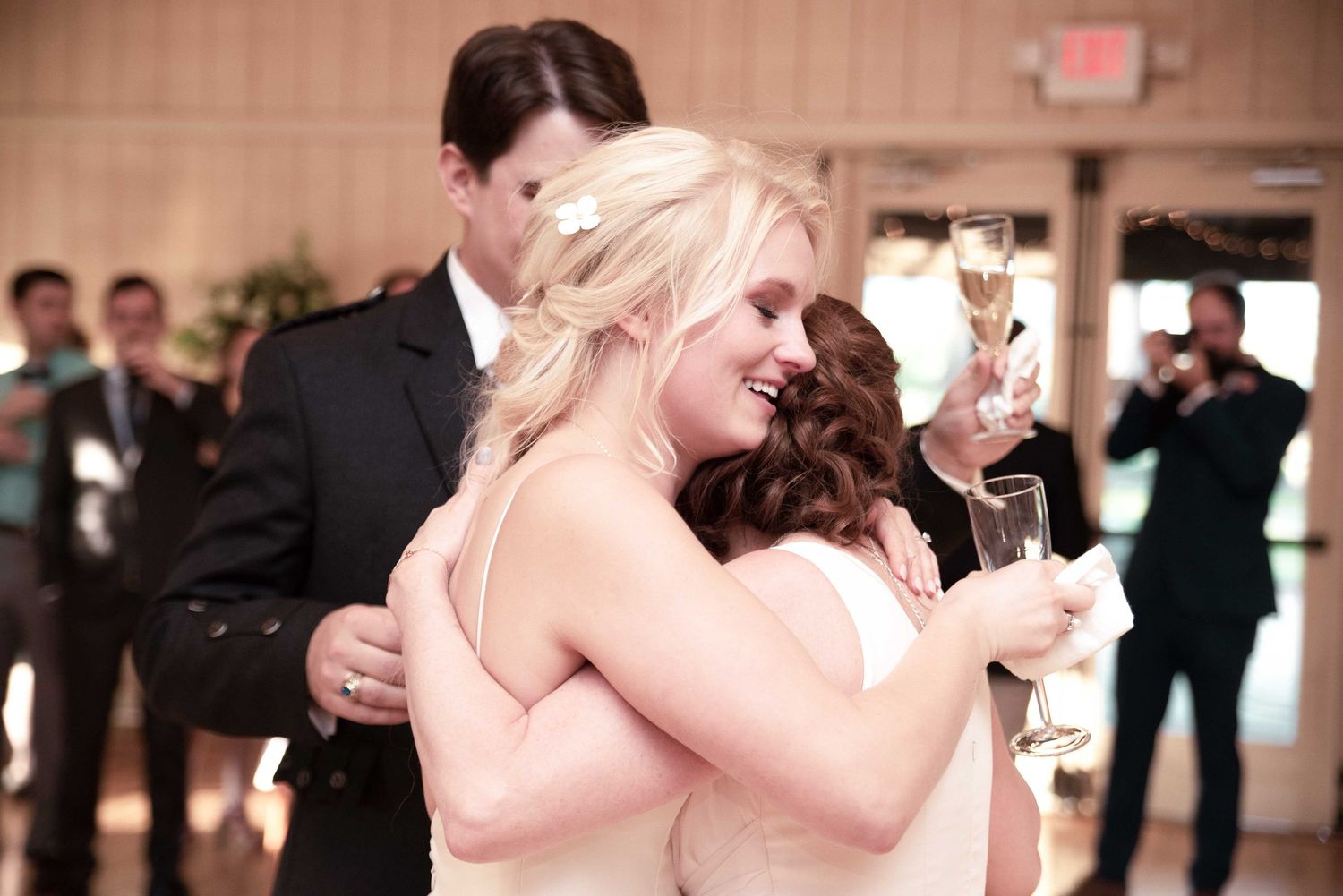 Two bridesmaids in white dresses share a warm hug while holding champagne glasses at a wedding reception.