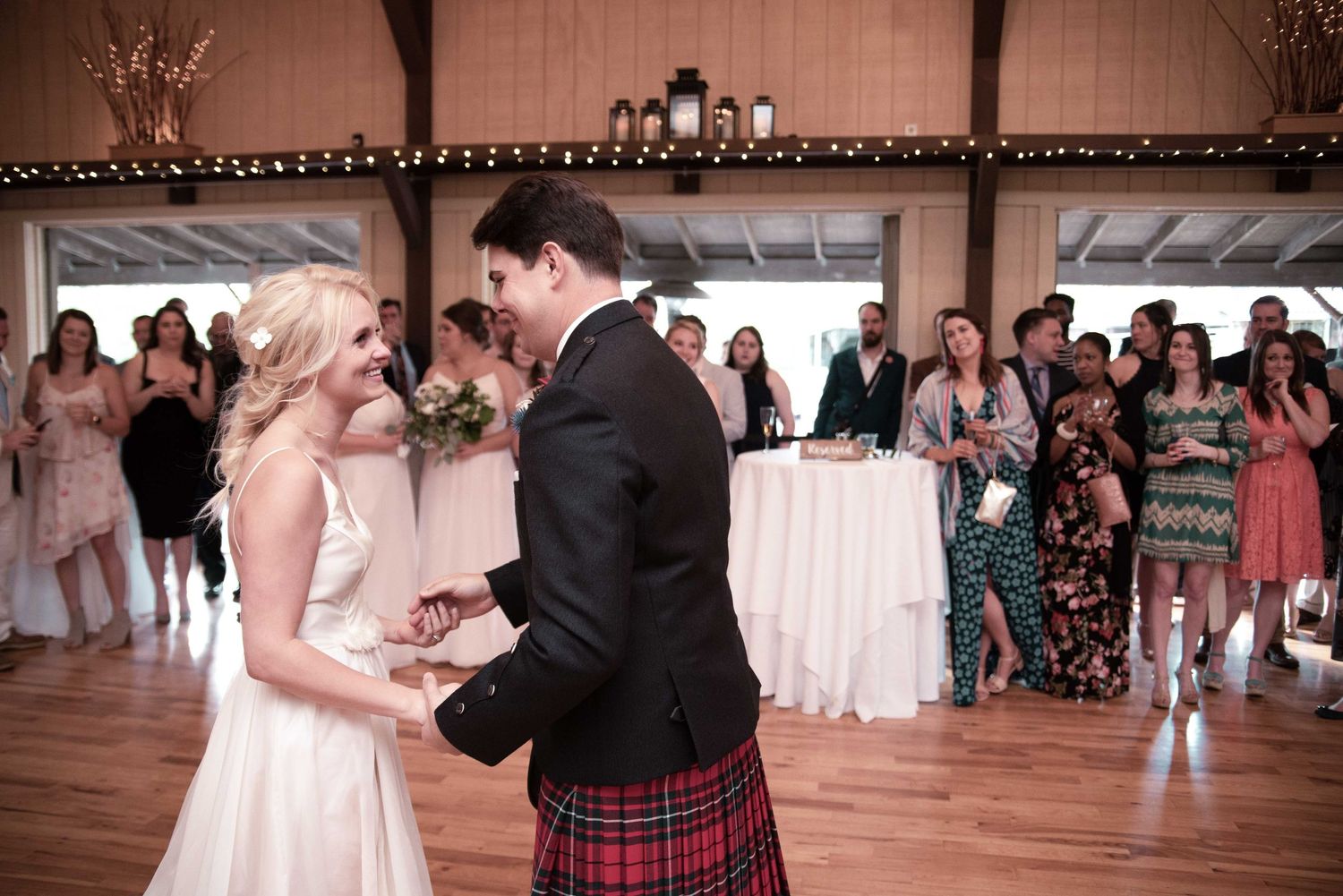 Bride and groom share first dance in rustic barn venue, groom wearing traditional Scottish tartan kilt.