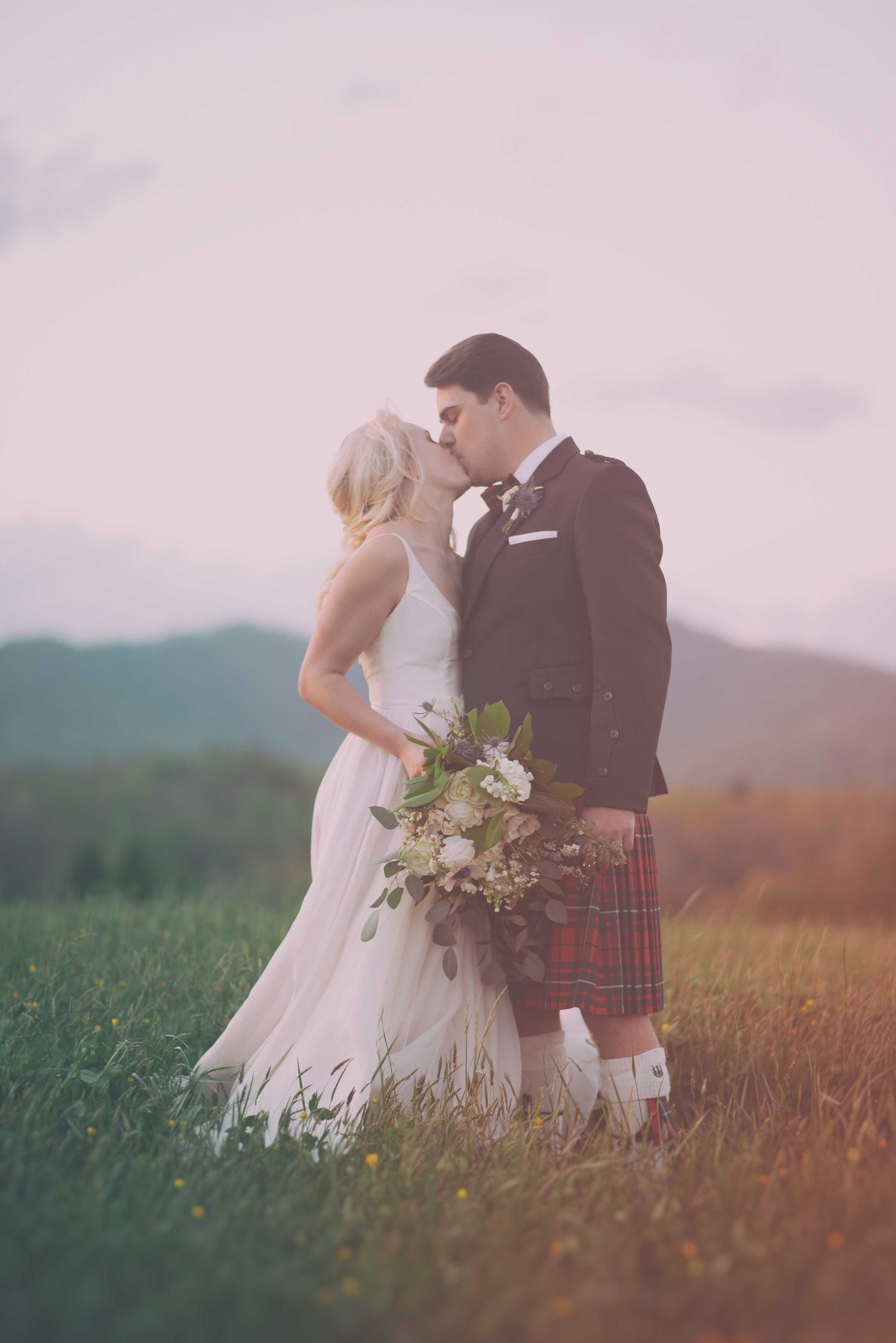 Bride and groom kissing in a green field at sunset, groom wearing a red tartan kilt and bride holding a bouquet.