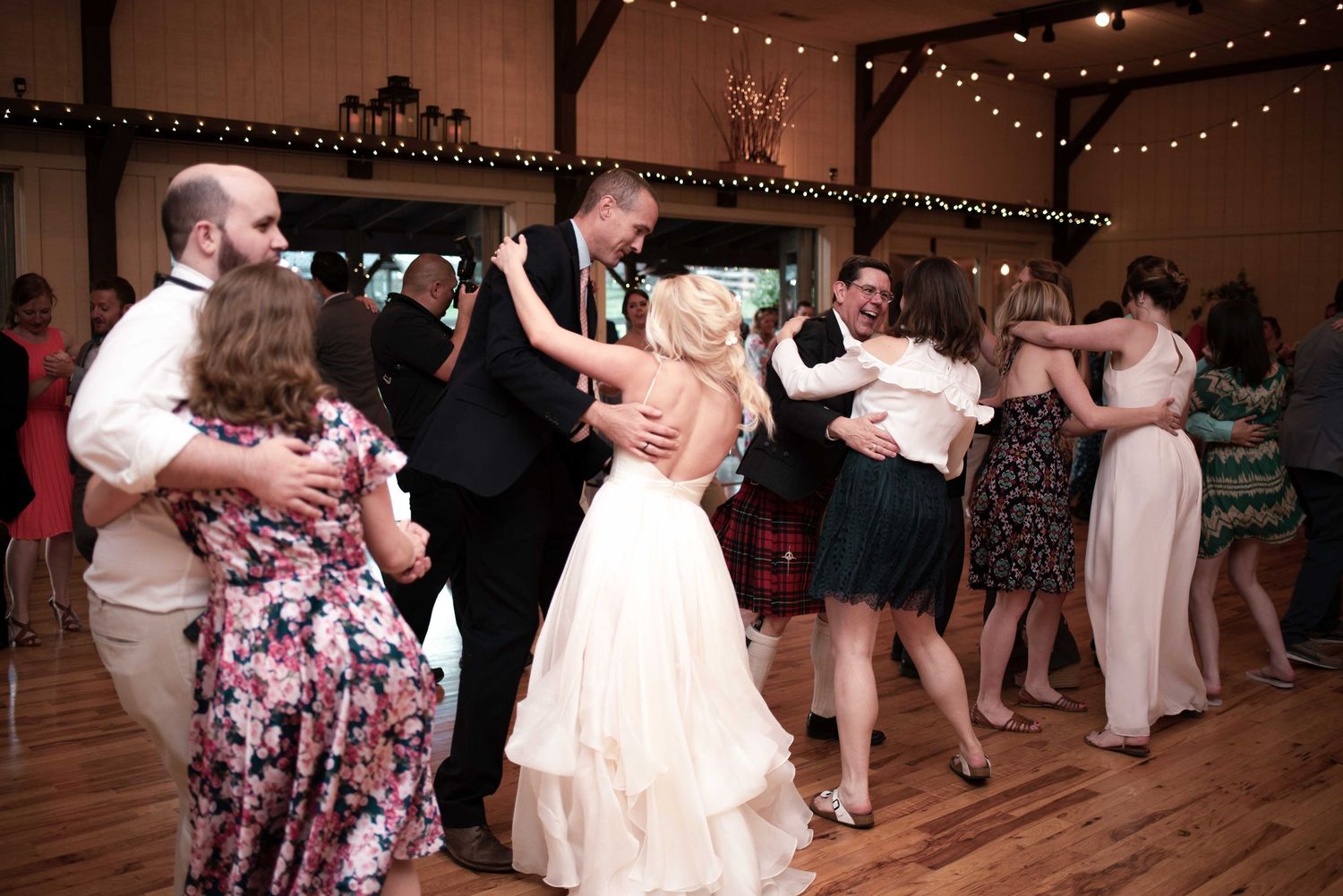 Bride and groom share first dance surrounded by guests at a rustic barn wedding reception with string lights.