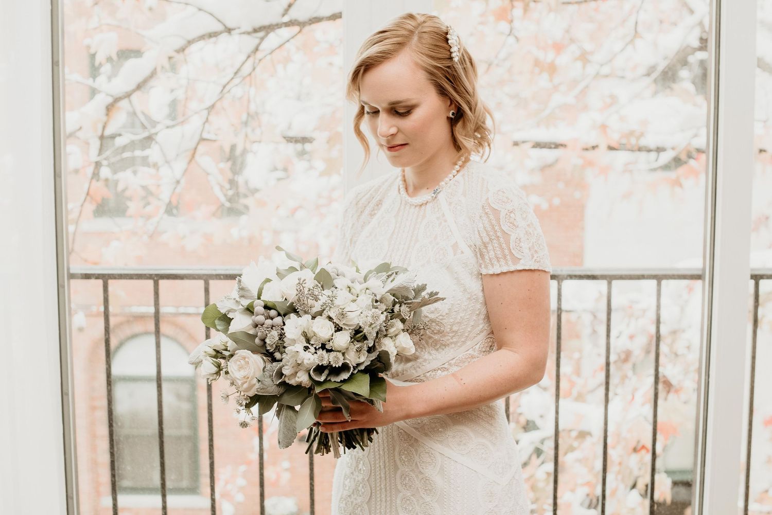 Bride in beaded white gown holding lush white and green bouquet on balcony with autumn trees in background.