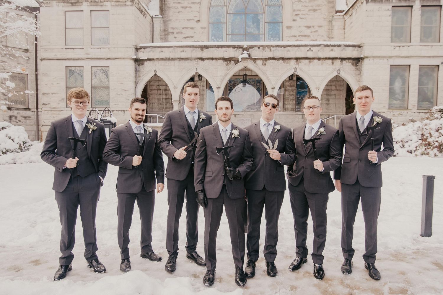 Groomsmen in dark suits pose outside a snowy stone church in a winter wedding party photo.