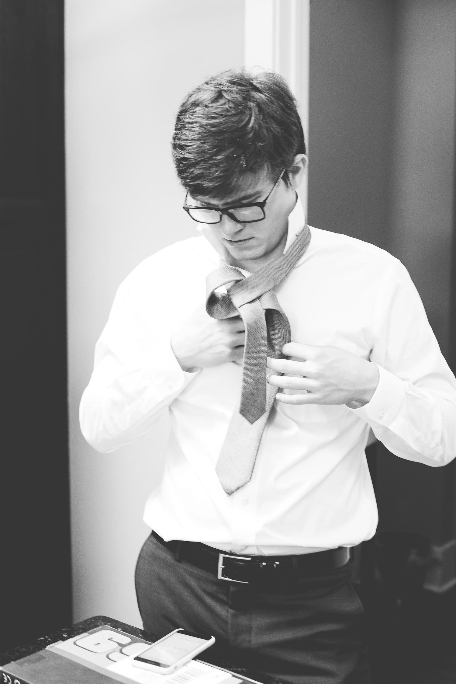 Black and white photo of a young man in glasses adjusting his tie while wearing a white dress shirt and dark trousers.