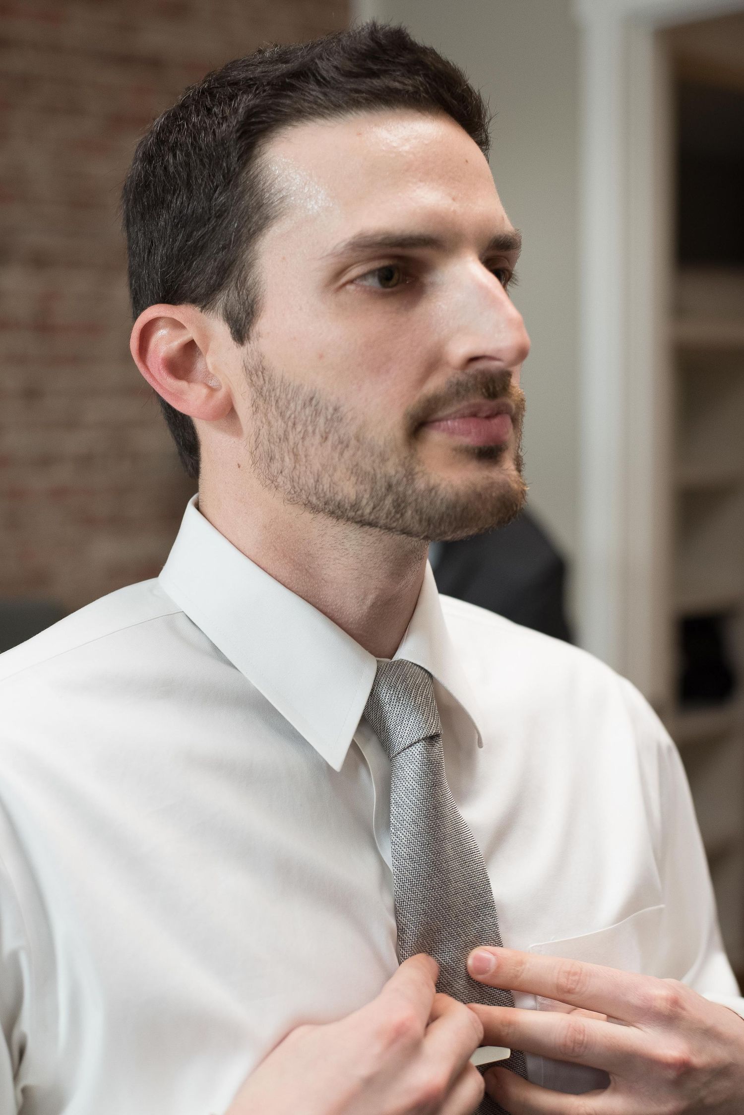 A young man with stubble adjusts his gray tie while wearing a white dress shirt in an indoor setting.