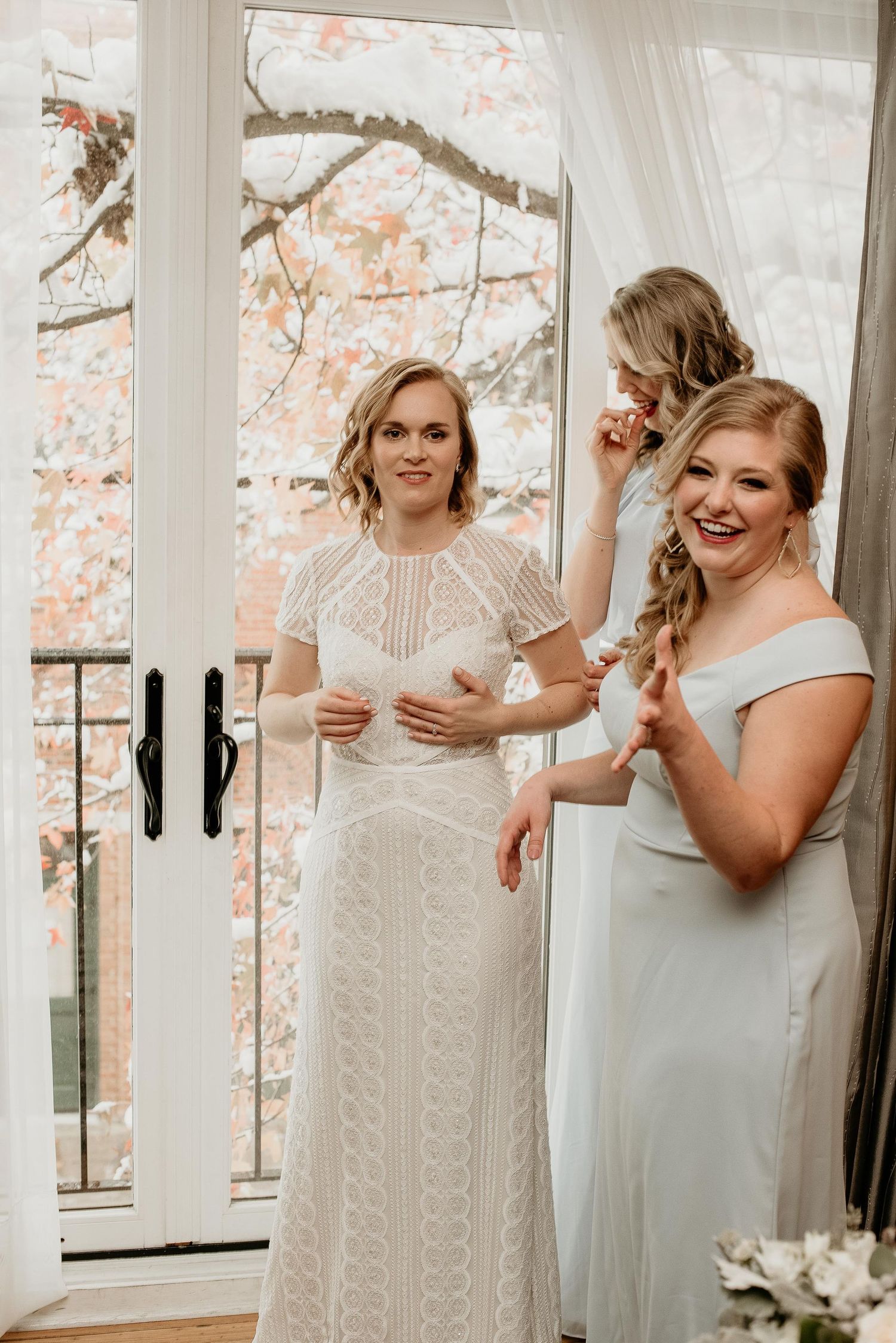 Bride in lace gown with bridesmaids in white dresses preparing near French doors with autumn trees in background.