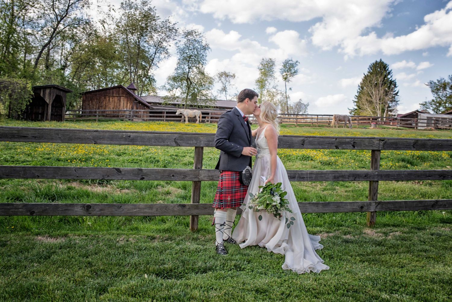 Bride and groom kissing by a rustic wooden fence on a green farm, groom wearing a traditional Scottish kilt.